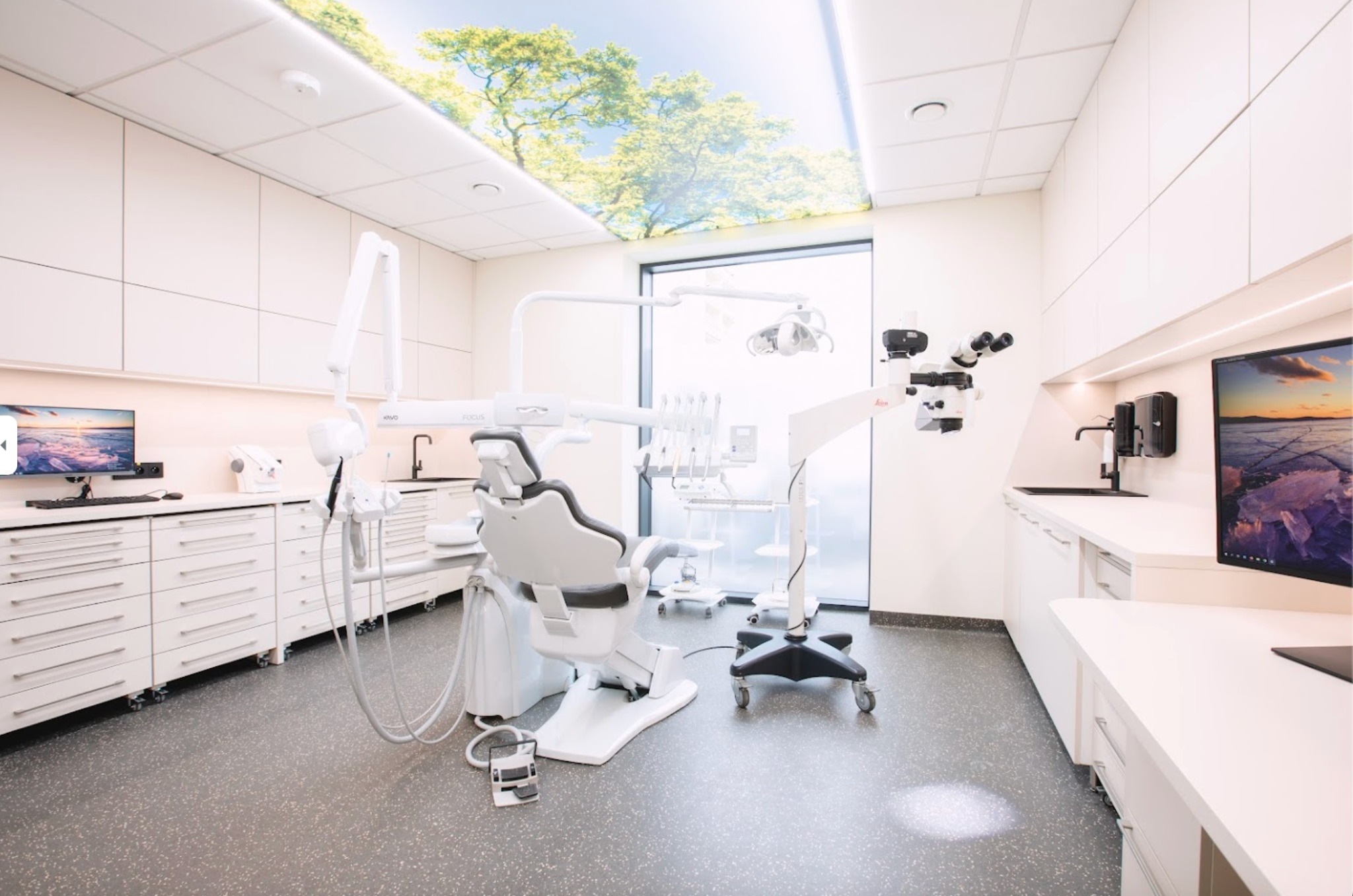 Nowoczesny gabinet stomatologiczny z białymi meblami, specjalistycznym sprzętem, mikroskopem Leica i fototapetą z motywem drzew na suficie, zapewniającą relaksującą atmosferę.