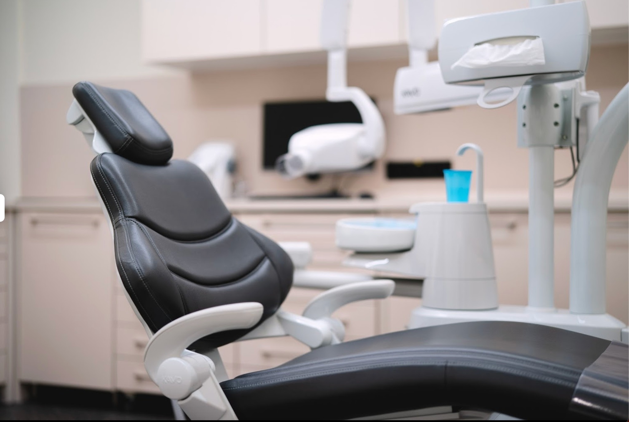 Czarne skórzane krzesło dentystyczne z białymi podłokietnikami w nowoczesnym gabinecie stomatologicznym, z widocznym sprzętem dentystycznym i szafkami w tle.