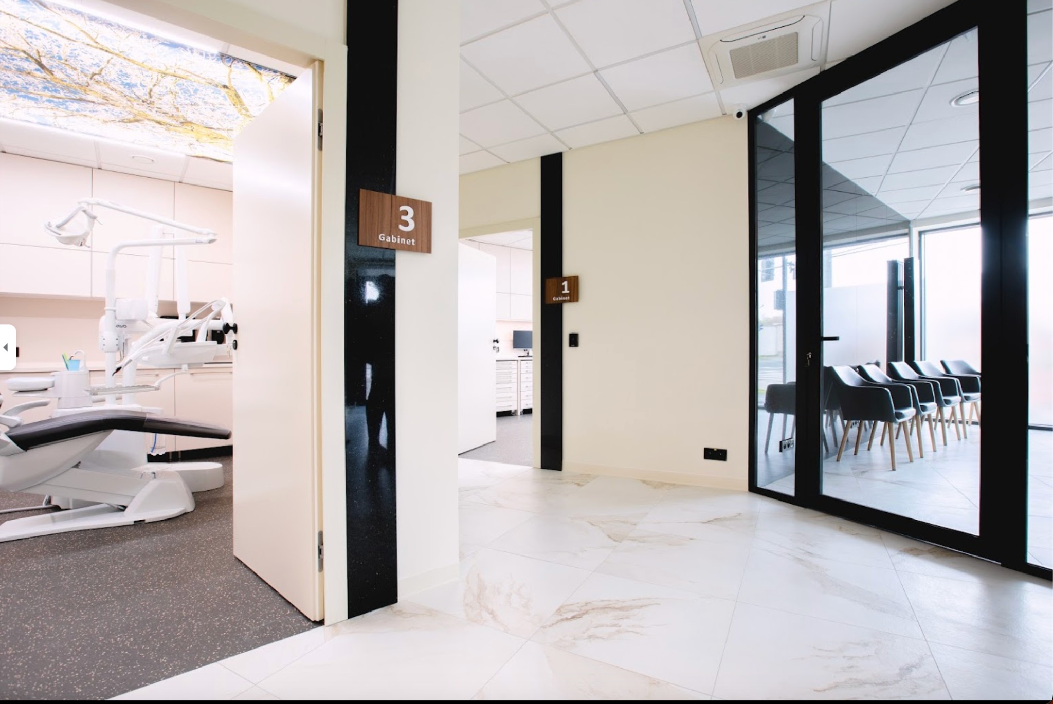 Nowoczesne wnętrze kliniki dentystycznej z marmurową podłogą, widok na gabinet z fotelem dentystycznym i poczekalnię z krzesłami.