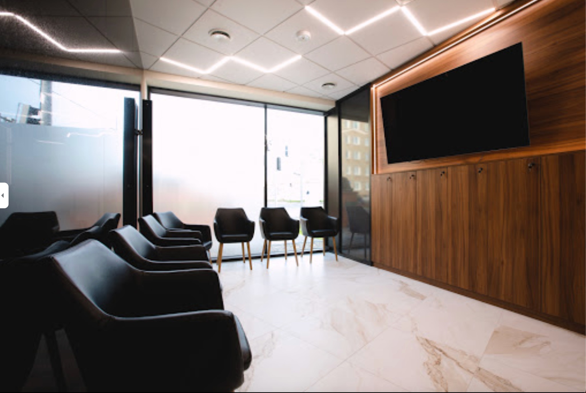 Nowoczesna poczekalnia z rzędem czarnych foteli, marmurową podłogą, drewnianą ścianą z wbudowanym telewizorem i geometrycznym oświetleniem sufitowym.