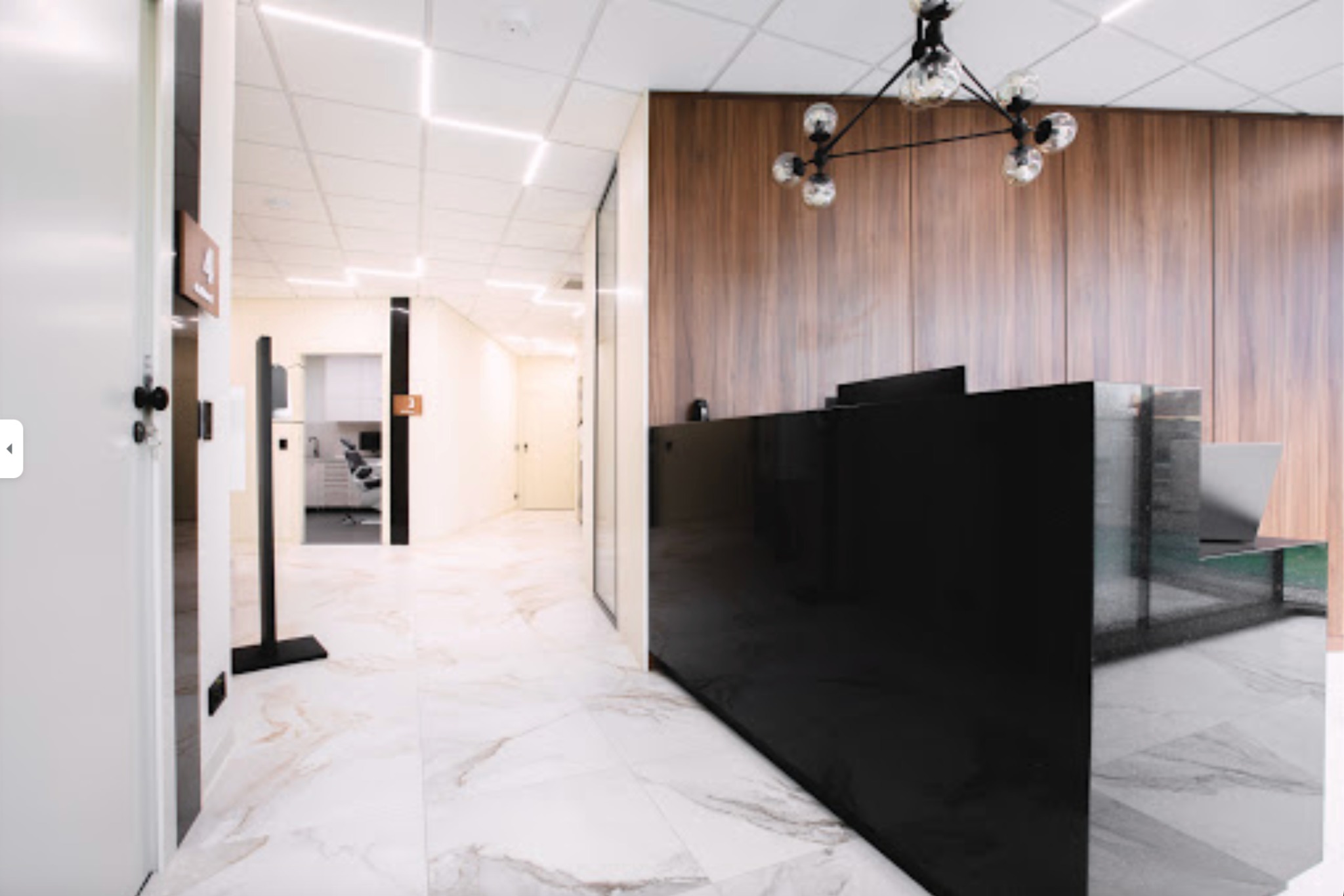 Nowoczesne wnętrze kliniki: recepcja z czarnym blatem, marmurowa podłoga i widok na gabinet z fotelem dentystycznym w tle.