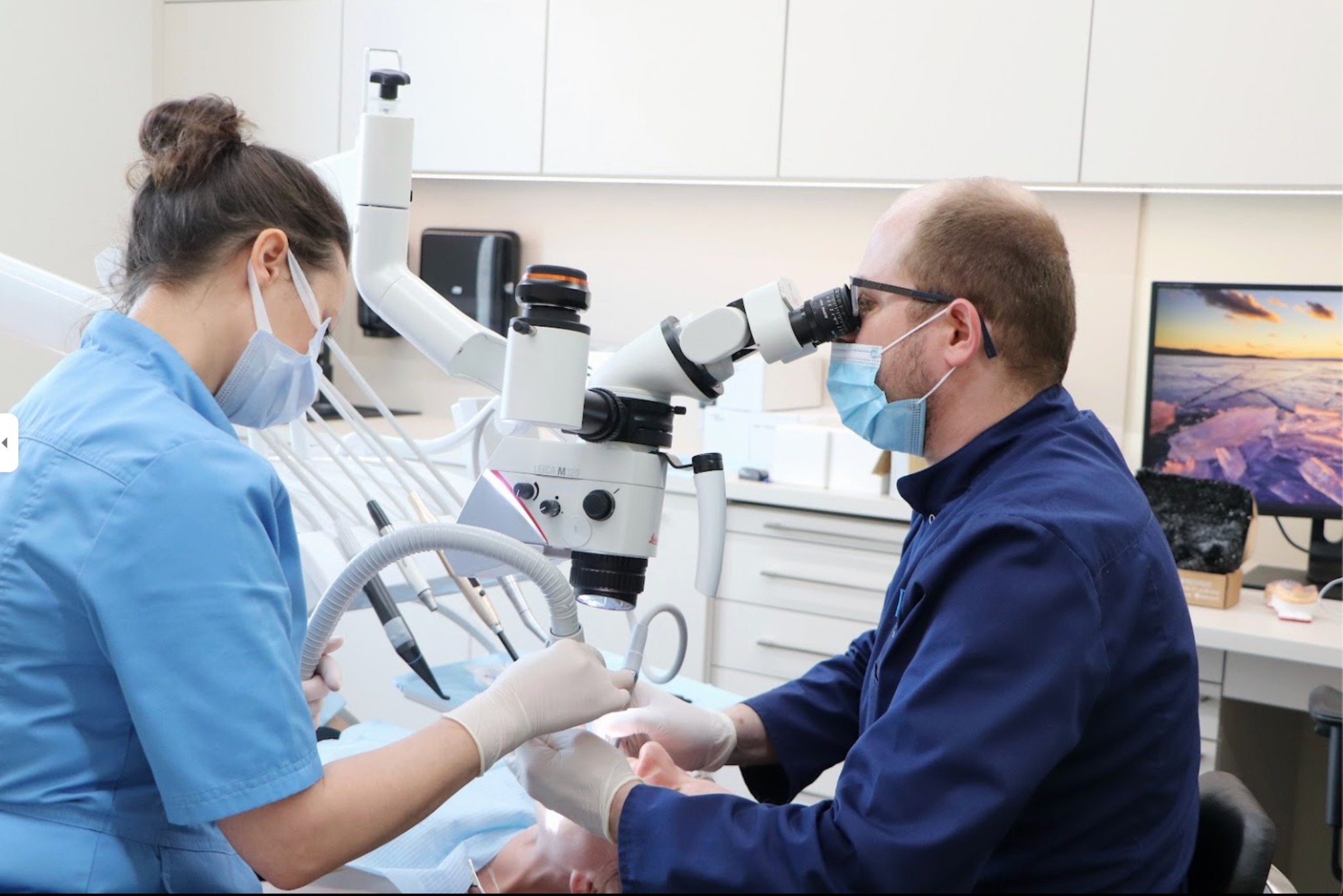 Lekarz dentysta w okularach i maseczce, przy użyciu mikroskopu stomatologicznego Leica M320, wykonuje zabieg w asyście pielęgniarki w nowoczesnym gabinecie.