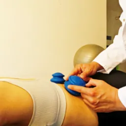 Masażysta w białym uniformie aplikuje niebieskie silikonowe bańki na plecy pacjentki leżącej na leżance, w tle widoczna złota piłka do ćwiczeń.