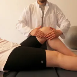 Pacjentka leży na kozetce, terapeuta w białym fartuchu wykonuje mobilizację stawu kolanowego, w tle widoczna szara piłka do ćwiczeń.