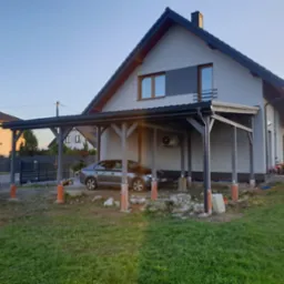 Nowoczesna, szara wiata samochodowa przylegająca do domu jednorodzinnego, wsparta na drewnianych słupach, z zaparkowanym szarym samochodem osobowym wewnątrz, widoczne prace wykończeniowe wokół wiaty.