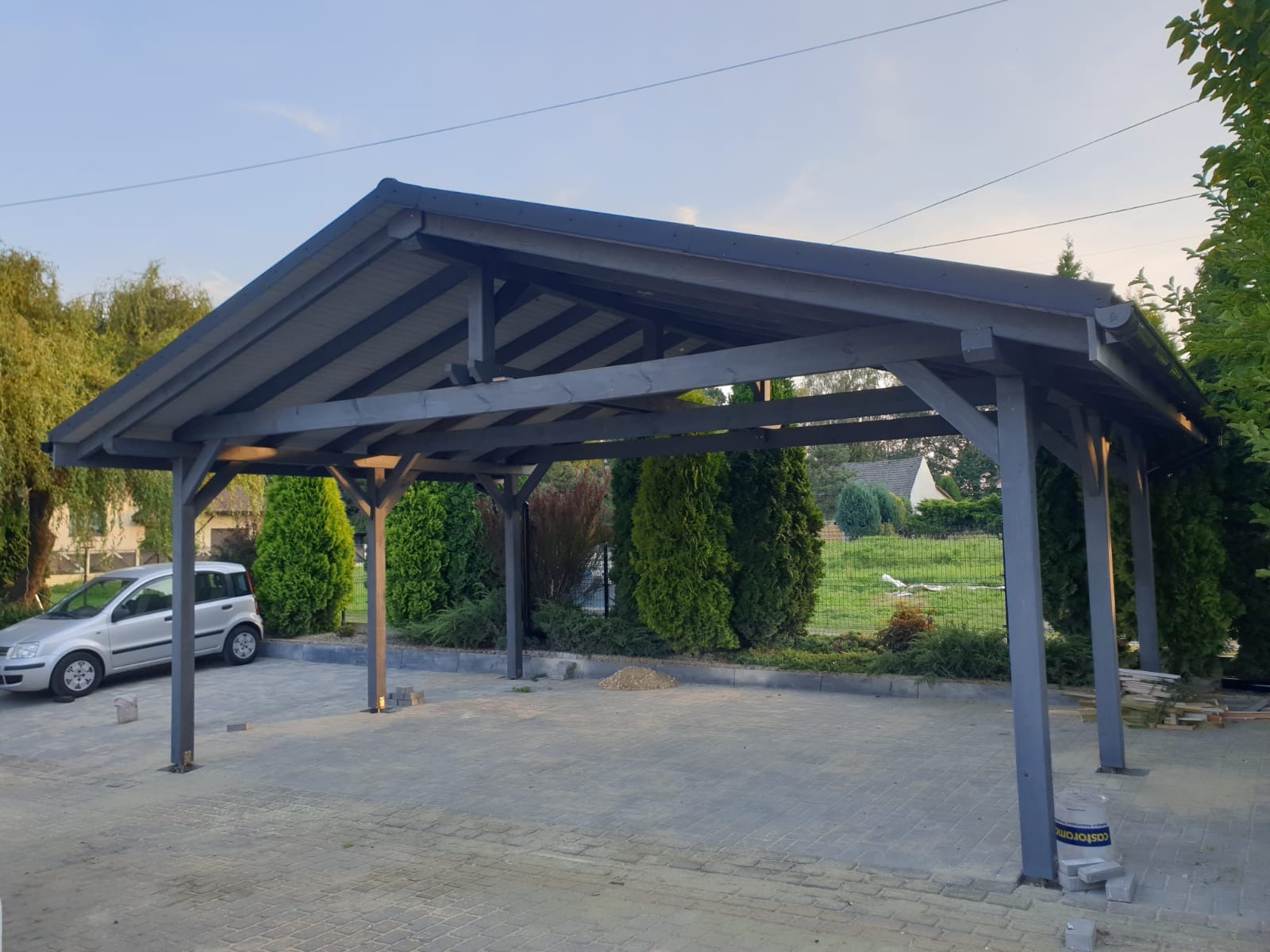 Szara, drewniana wiata garażowa z ukośnym dachem, widok z zewnątrz, obok zaparkowany srebrny samochód, kostka brukowa na podjeździe, w tle zielone tuje i dom.