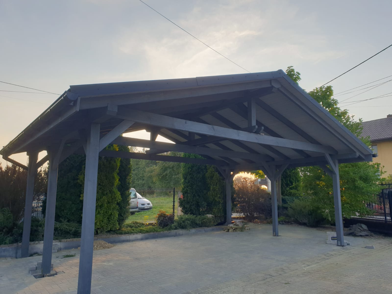 Drewniana wiata garażowa w odcieniach szarości, z widoczną konstrukcją dachu i słupami, na tle zieleni i błękitnego nieba o zmierzchu. Na podjeździe ułożone kostki brukowe.