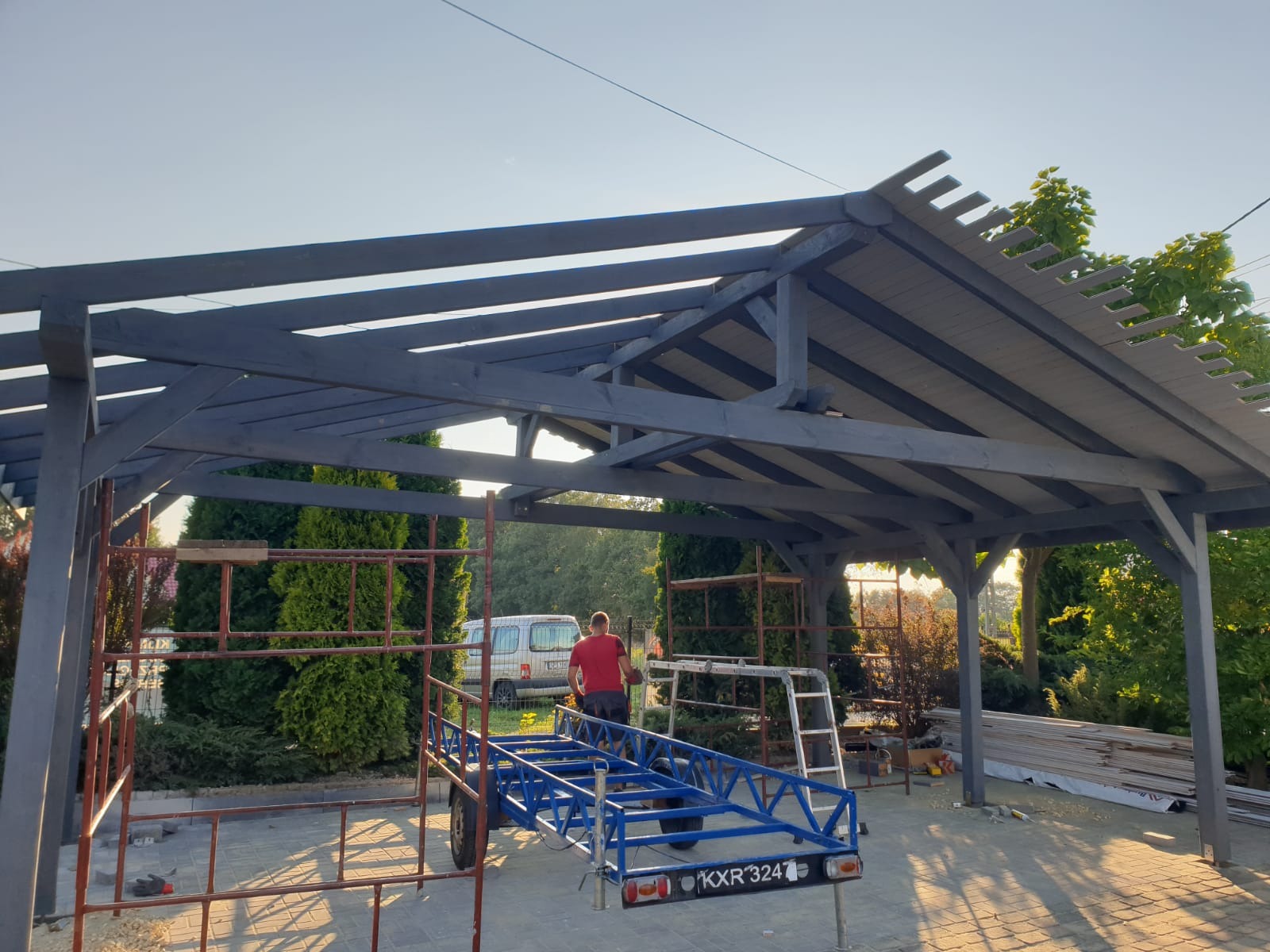 Konstrukcja altany w trakcie budowy, widoczne rusztowania, przyczepka, częściowo zadaszona, pomalowana na szaro, w tle zieleń i zaparkowany samochód.