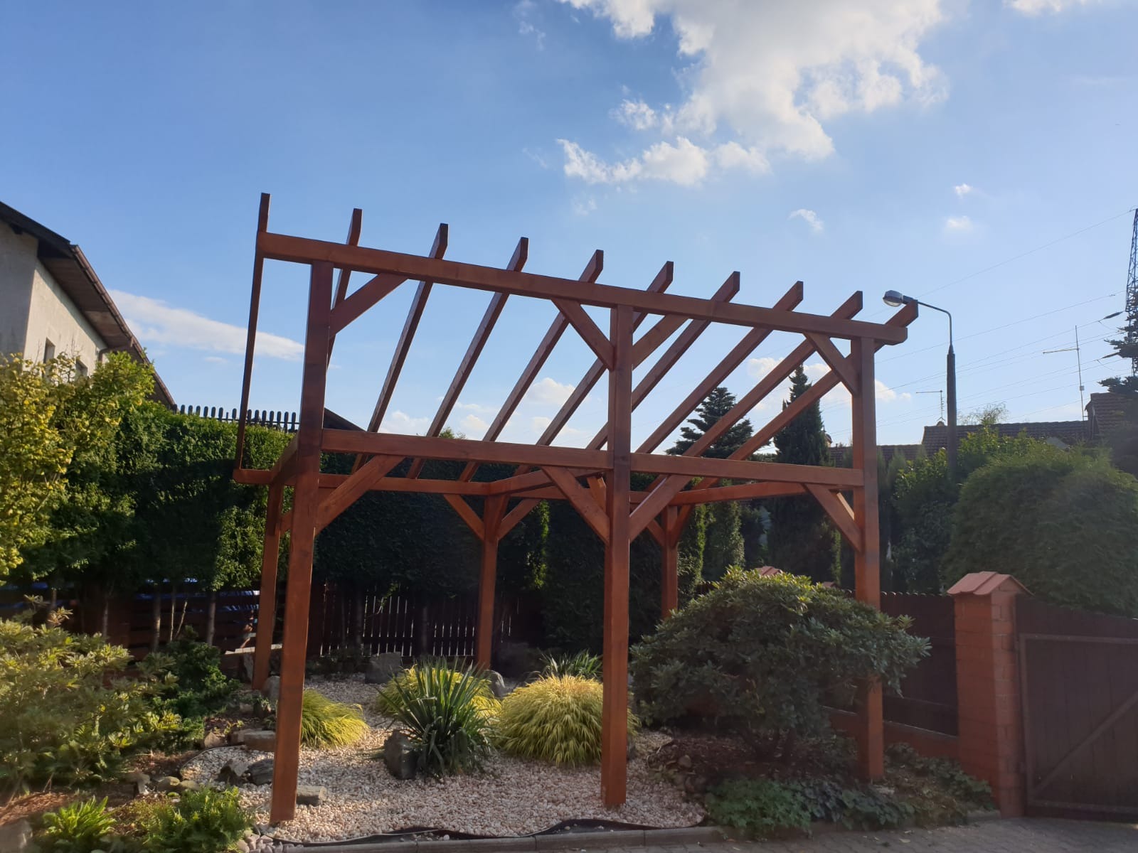 Drewniana konstrukcja altany w ogrodzie, widoczne elementy nośne i zadaszenie, otoczenie z krzewami i białym żwirem, błękitne niebo w tle.