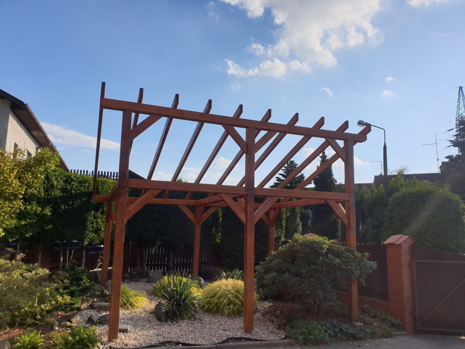 Surowa, drewniana konstrukcja altany w trakcie budowy w Pszczynie, otoczona dekoracyjnym ogrodem z białym żwirem i roślinami.