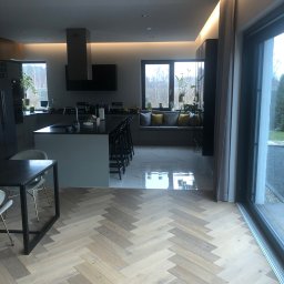 Gracz development sp zoo - Jasny parkiet ułożony w jodełkę francuską w przestronnej kuchni z wyspą, marmurowa podłoga w tle, duże okno przesuwne.