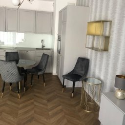 Gracz development sp zoo - Jasnoszara kuchnia z parkietem w jodełkę, okrągłym stołem i aksamitnymi krzesłami, złote detale lampy i półki.