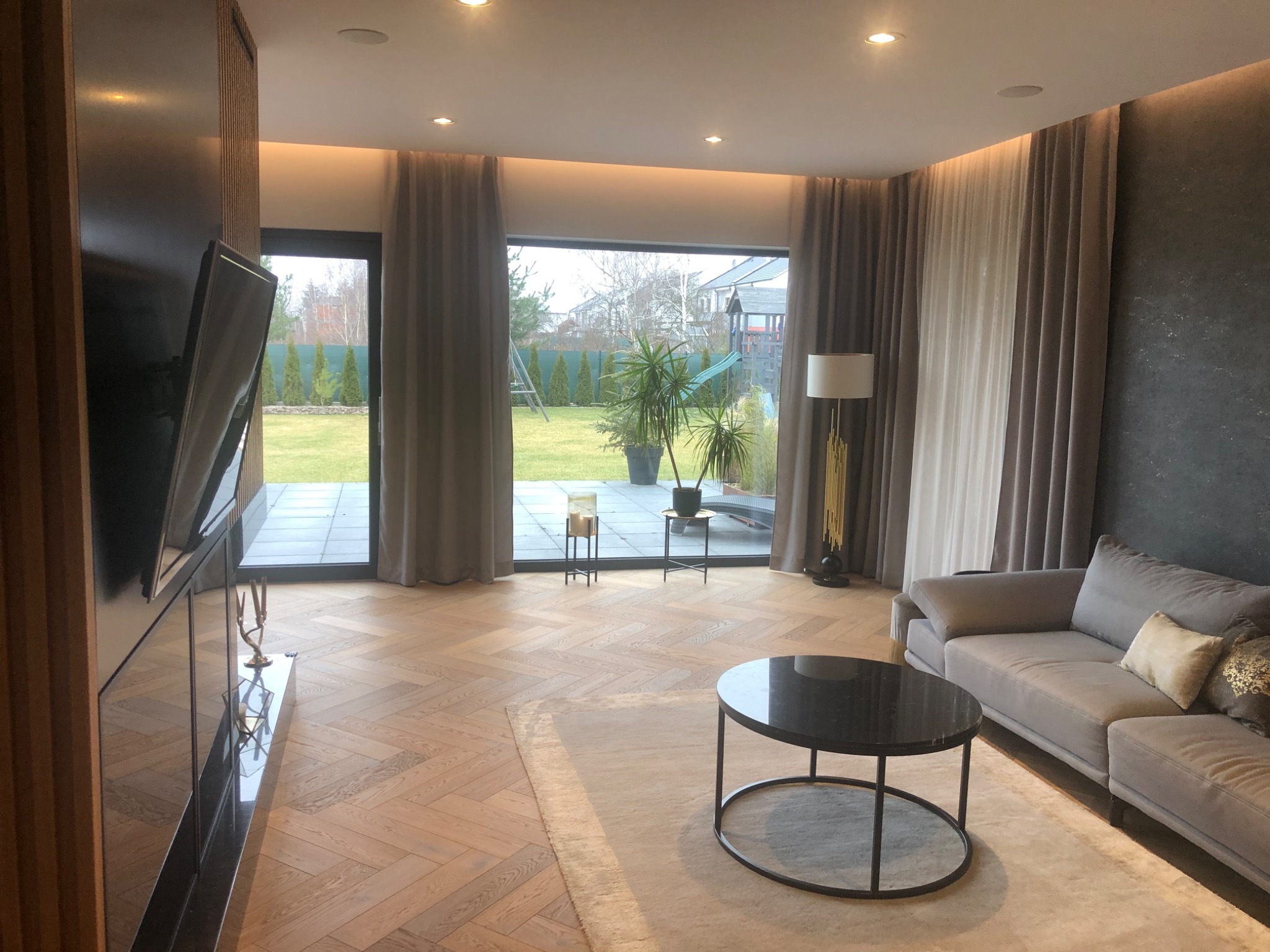 Jasny salon z parkietem w jodełkę, szarą sofą i widokiem na ogród przez duże okno z zasłonami.