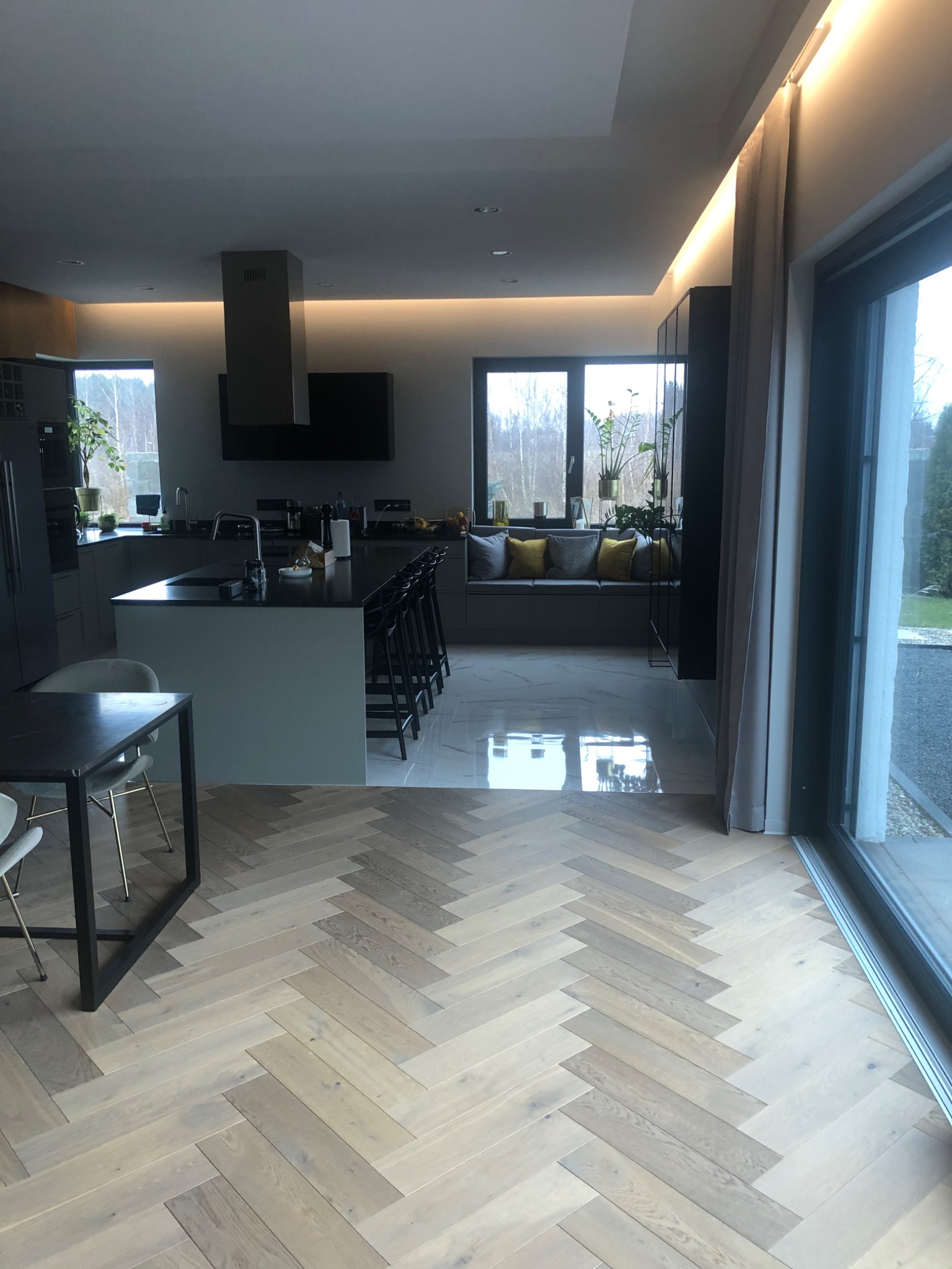 Jasny parkiet ułożony w jodełkę francuską w przestronnej kuchni z wyspą, marmurowa podłoga w tle, duże okno przesuwne.