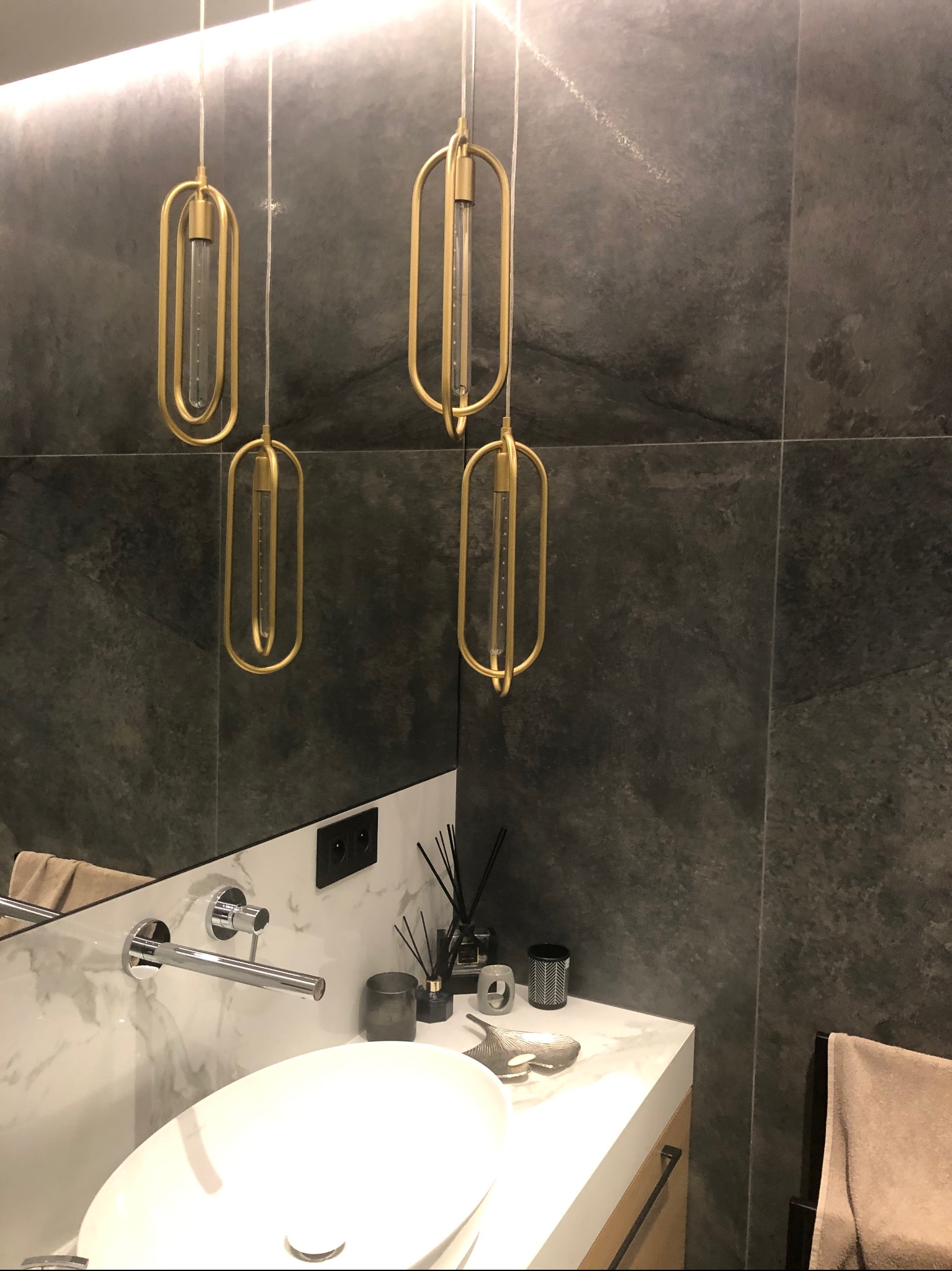 Nowoczesna łazienka z umywalką nablatową, armaturą chromowaną i czterema wiszącymi lampami o złotych, podłużnych oprawach na tle ściany z ciemnoszarej płytki imitującej kamień.