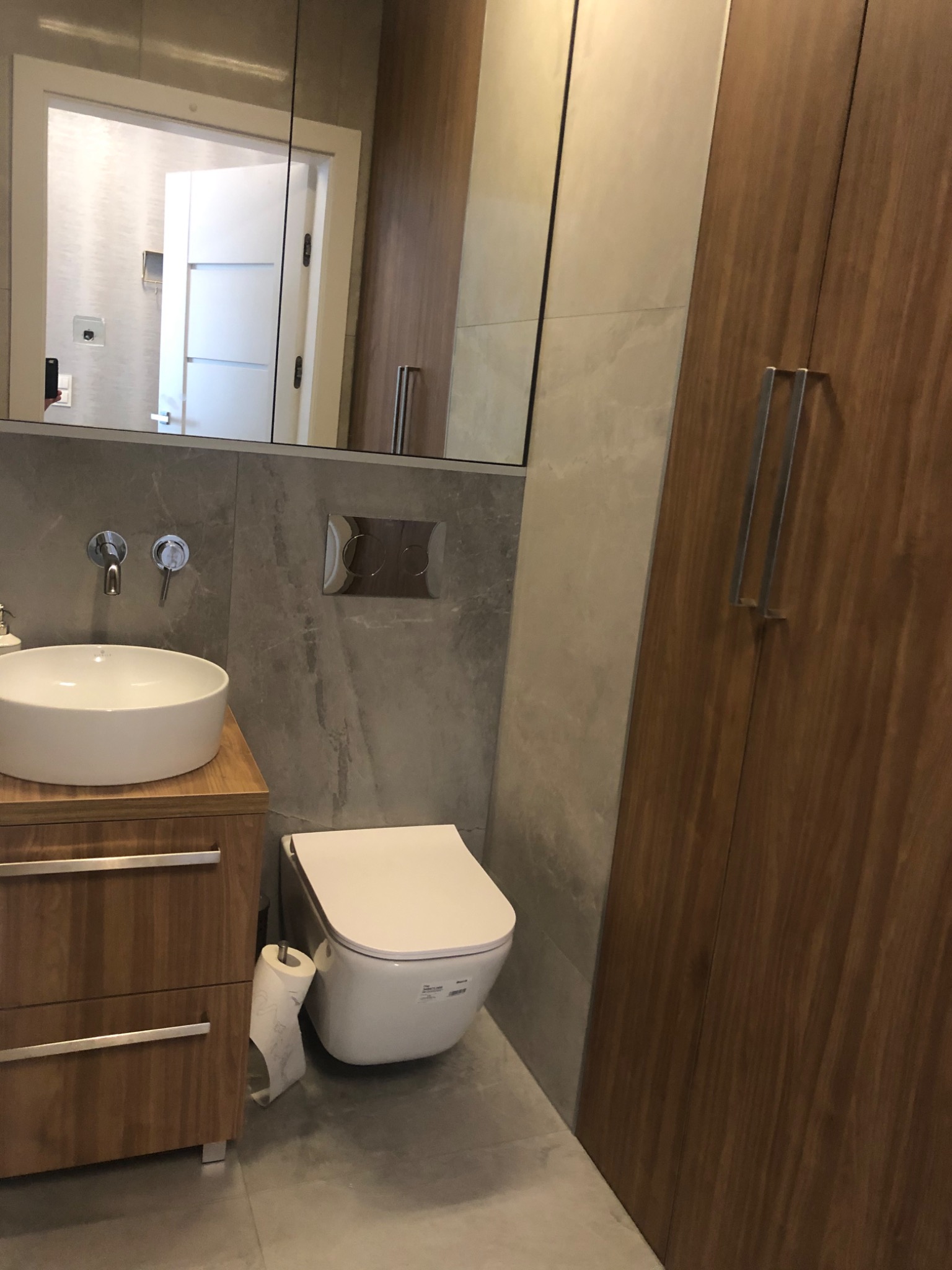 Nowoczesna łazienka z umywalką nablatową na drewnianej szafce, podwieszaną toaletą i szarą płytką imitującą kamień.