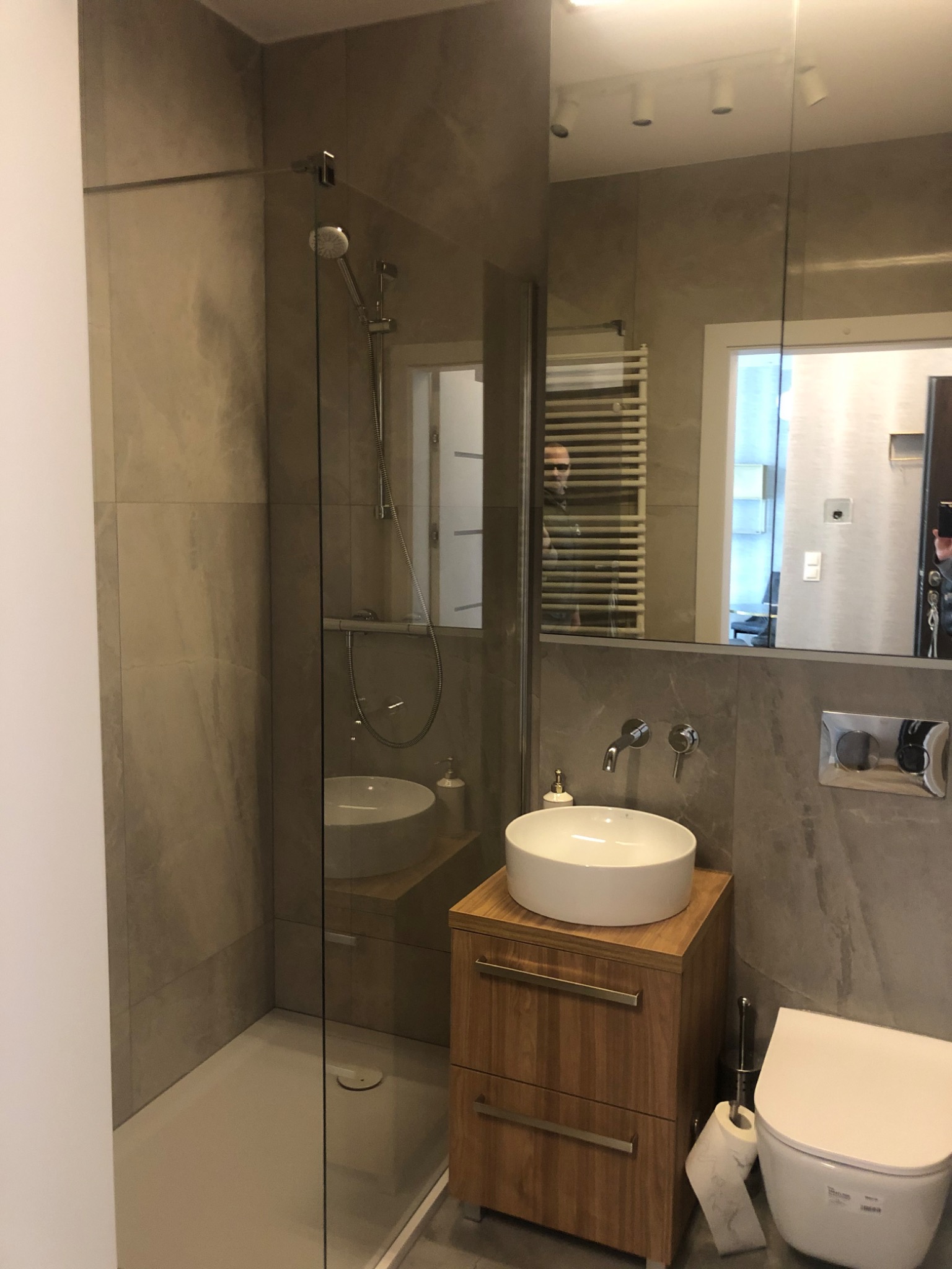 Nowoczesna łazienka z kabiną prysznicową typu walk-in, umywalką nablatową na drewnianej szafce z szufladami i wiszącą toaletą, wykończona szarymi płytkami imitującymi kamień.