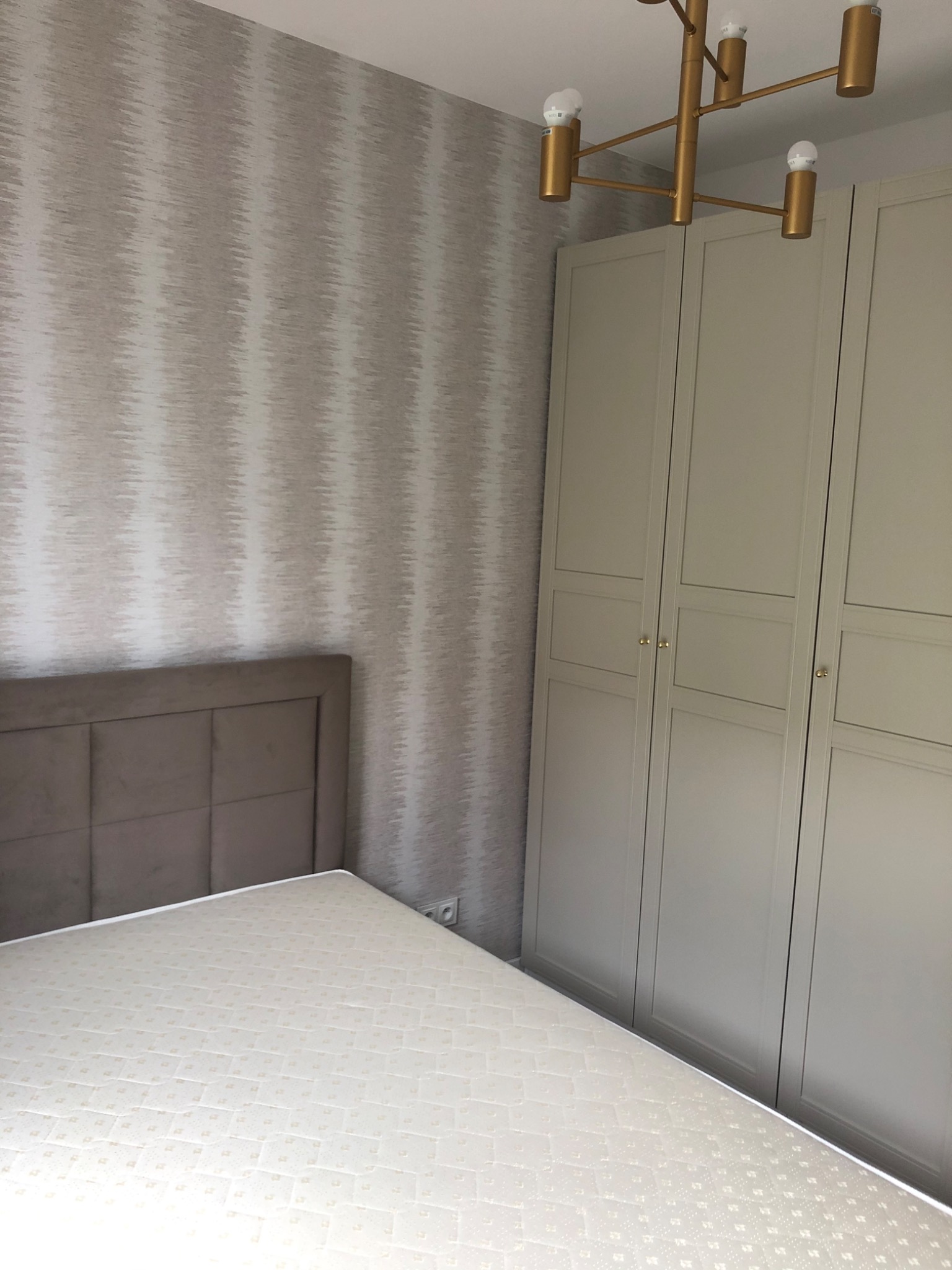 Sypialnia po remoncie: łóżko z beżowym wezgłowiem, wzorzysta tapeta w odcieniach szarości i beżu, oraz jasnoszara szafa trzydrzwiowa z delikatnymi frezami, oświetlona złotą lampą sufitową.