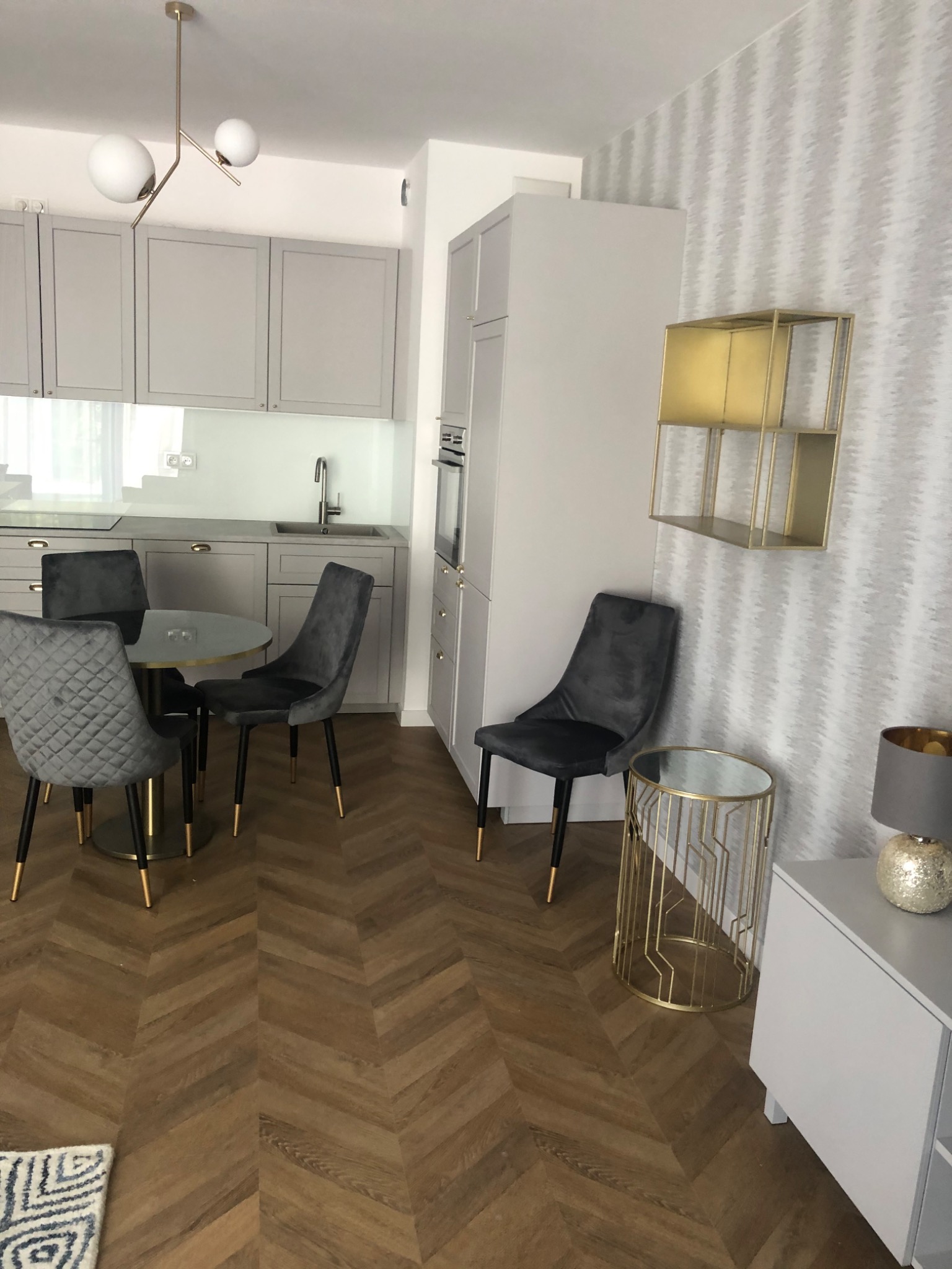 Jasnoszara kuchnia z parkietem w jodełkę, okrągłym stołem i aksamitnymi krzesłami, złote detale lampy i półki.