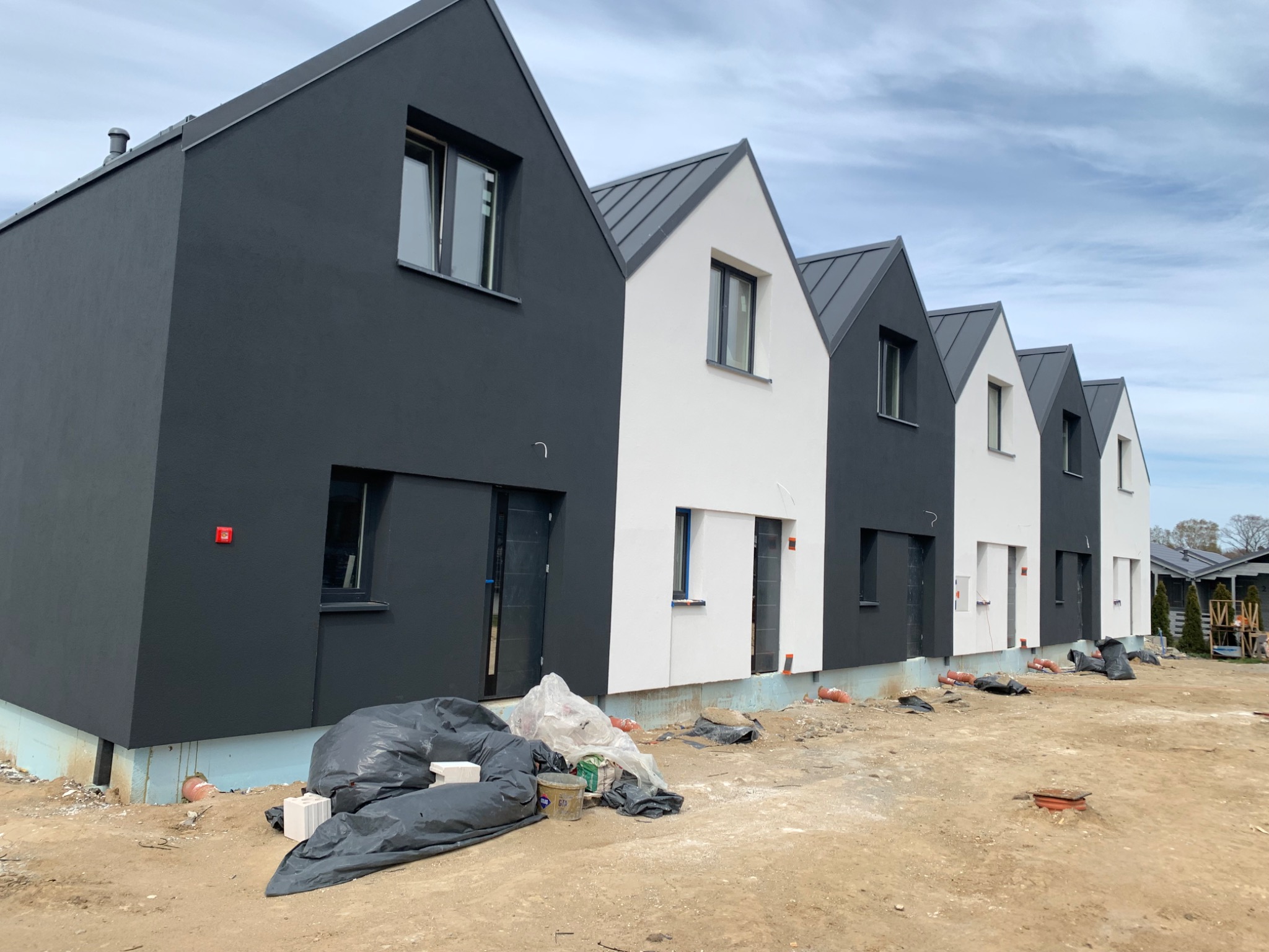 Rząd nowych domów jednorodzinnych w trakcie wykończenia, z czarno-białymi elewacjami, widoczne worki z materiałami budowlanymi i gruz na ziemi.