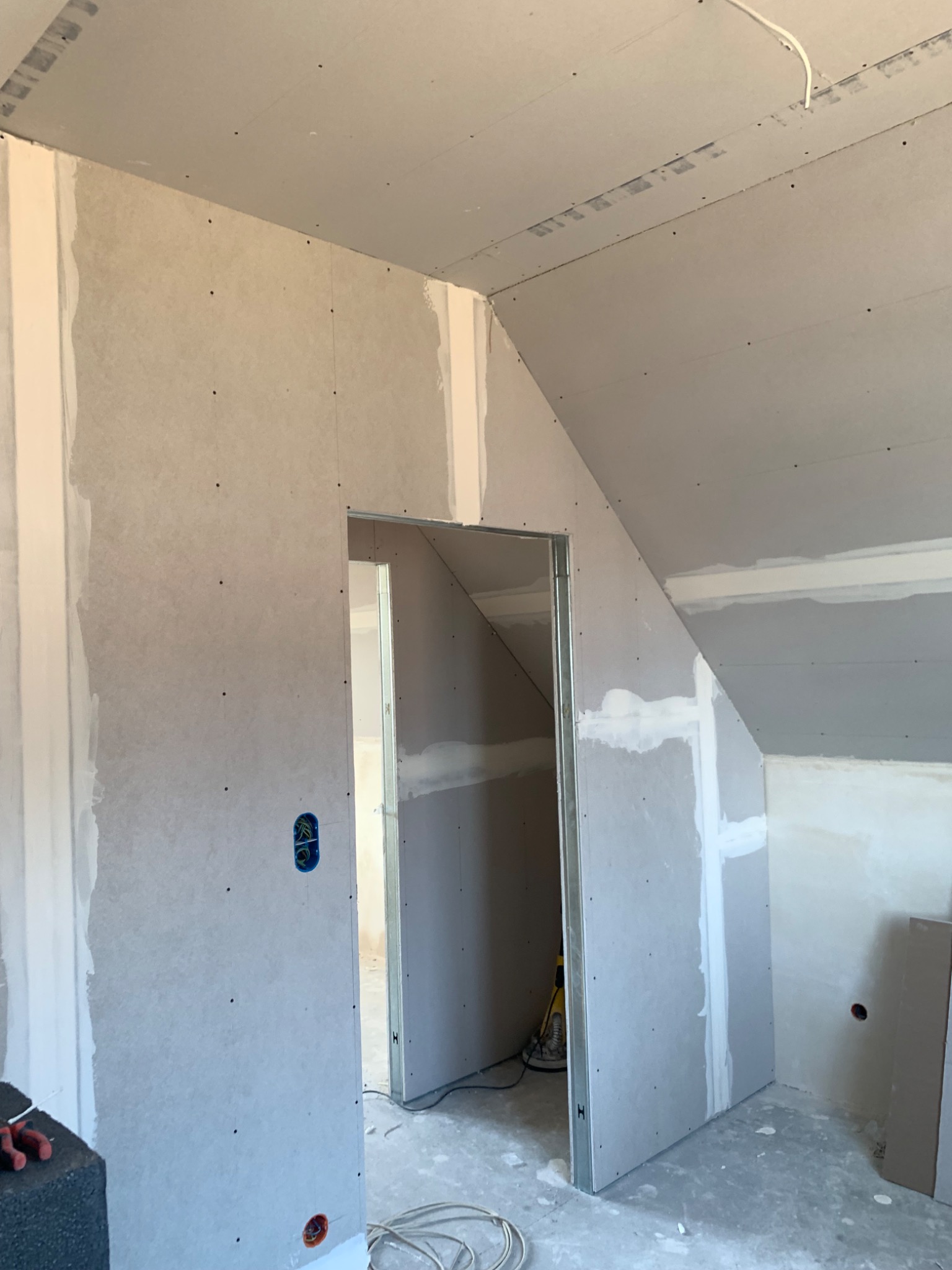 Montaż płyt karton gips na poddaszu ze skośnym sufitem, widoczne profile metalowe, szpachlowane łączenia i puszki elektryczne w ścianie.