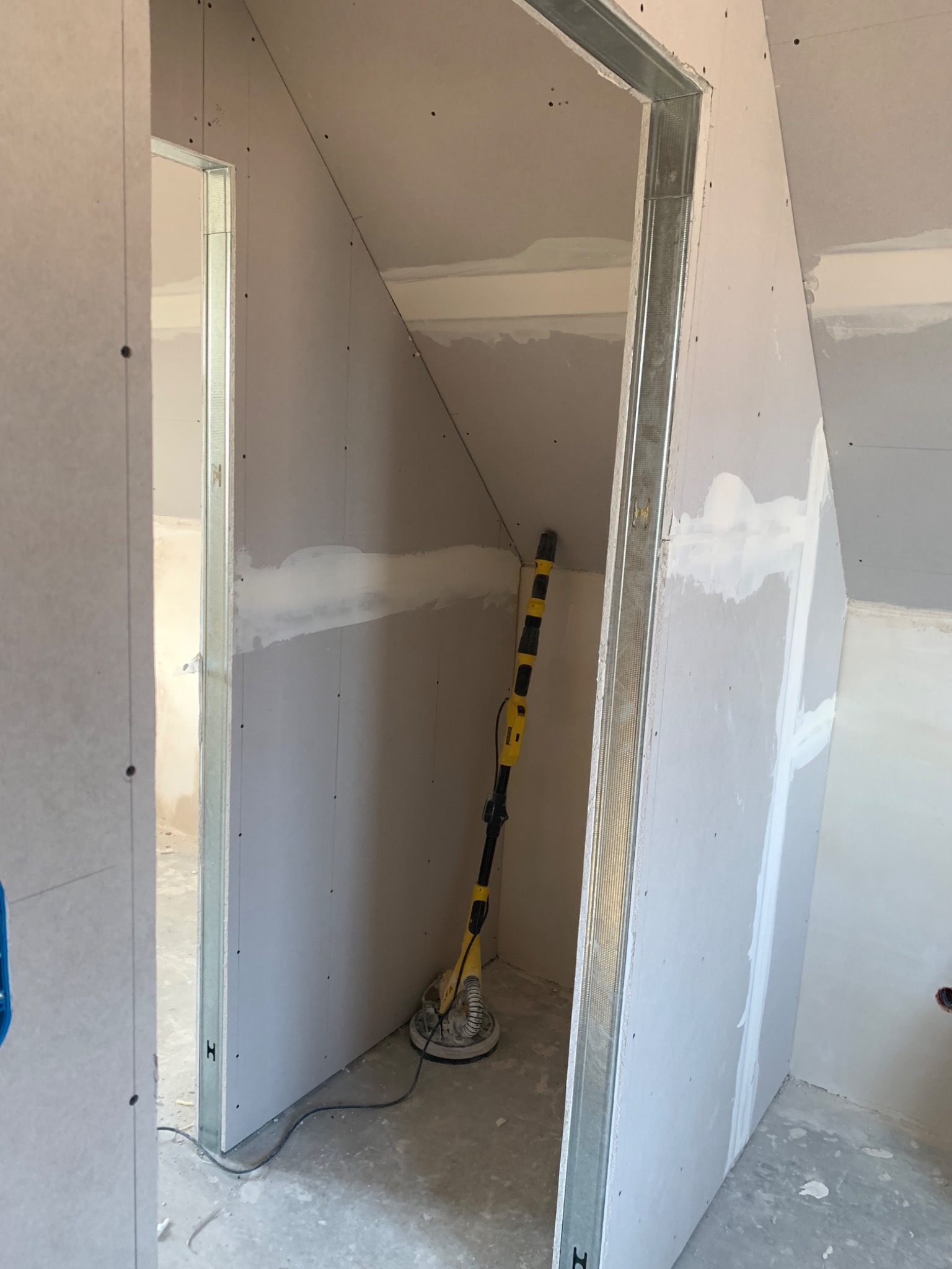 Wnętrze w trakcie prac wykończeniowych: ściany z płyt kartonowo-gipsowych, częściowo wyszpachlowane, z widoczną konstrukcją metalową ościeżnicy i szlifierką do gładzi na podłodze.