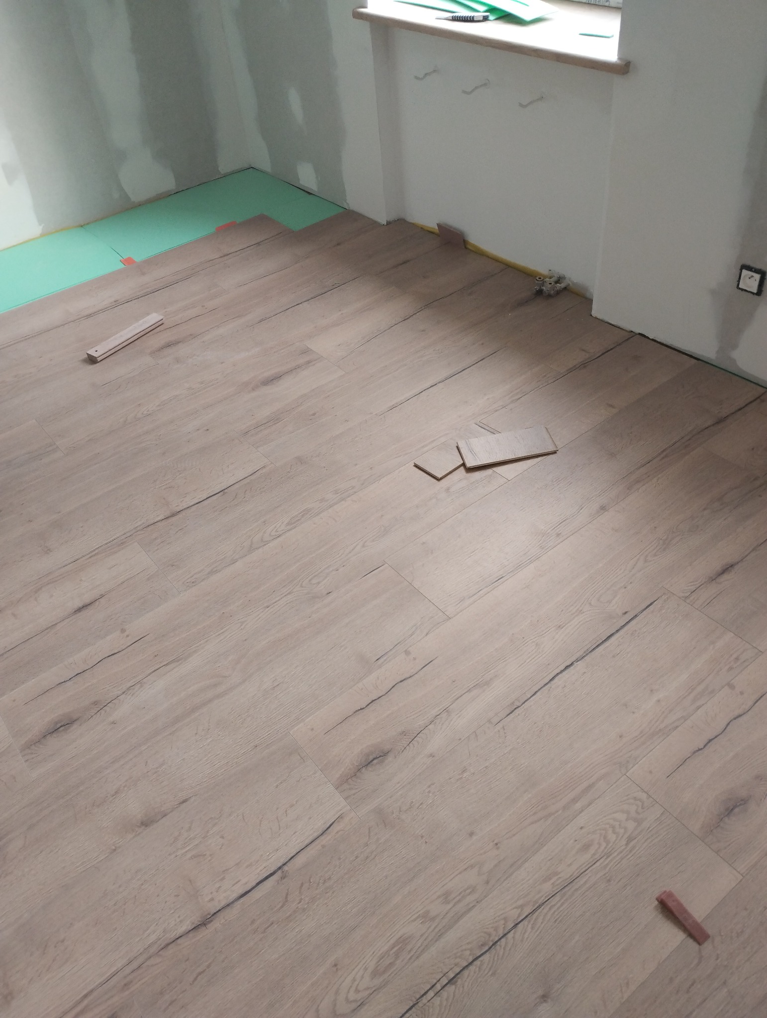 Fragment pokoju w trakcie montażu paneli podłogowych, widoczne podłoże izolacyjne, kilka ułożonych paneli o wzorze drewna i fragmenty paneli przy ścianie, ściany w stanie surowym.