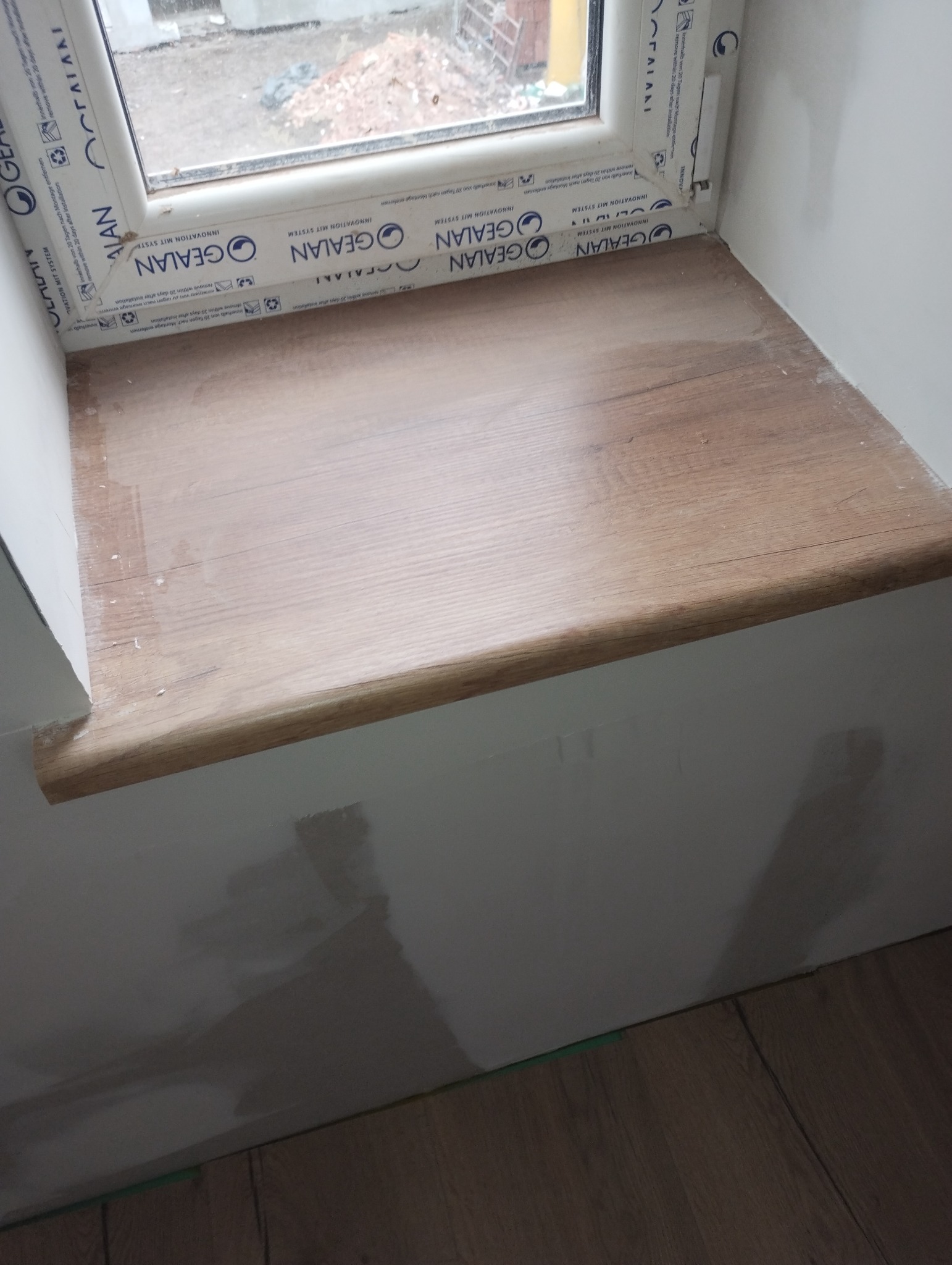 Parapet okienny z paneli drewnopodobnych, po montażu, z widocznymi śladami szpachlowania na ścianie i ramie okiennej.