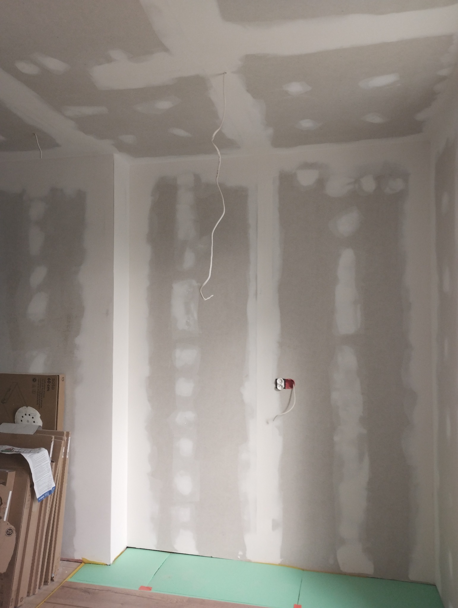 Pokój w trakcie prac wykończeniowych: ściany i sufit pokryte płytami kartonowo-gipsowymi, częściowo wyszpachlowane, widoczne kable elektryczne i gniazdko, na podłodze zielone płyty ochronne, w rogu...