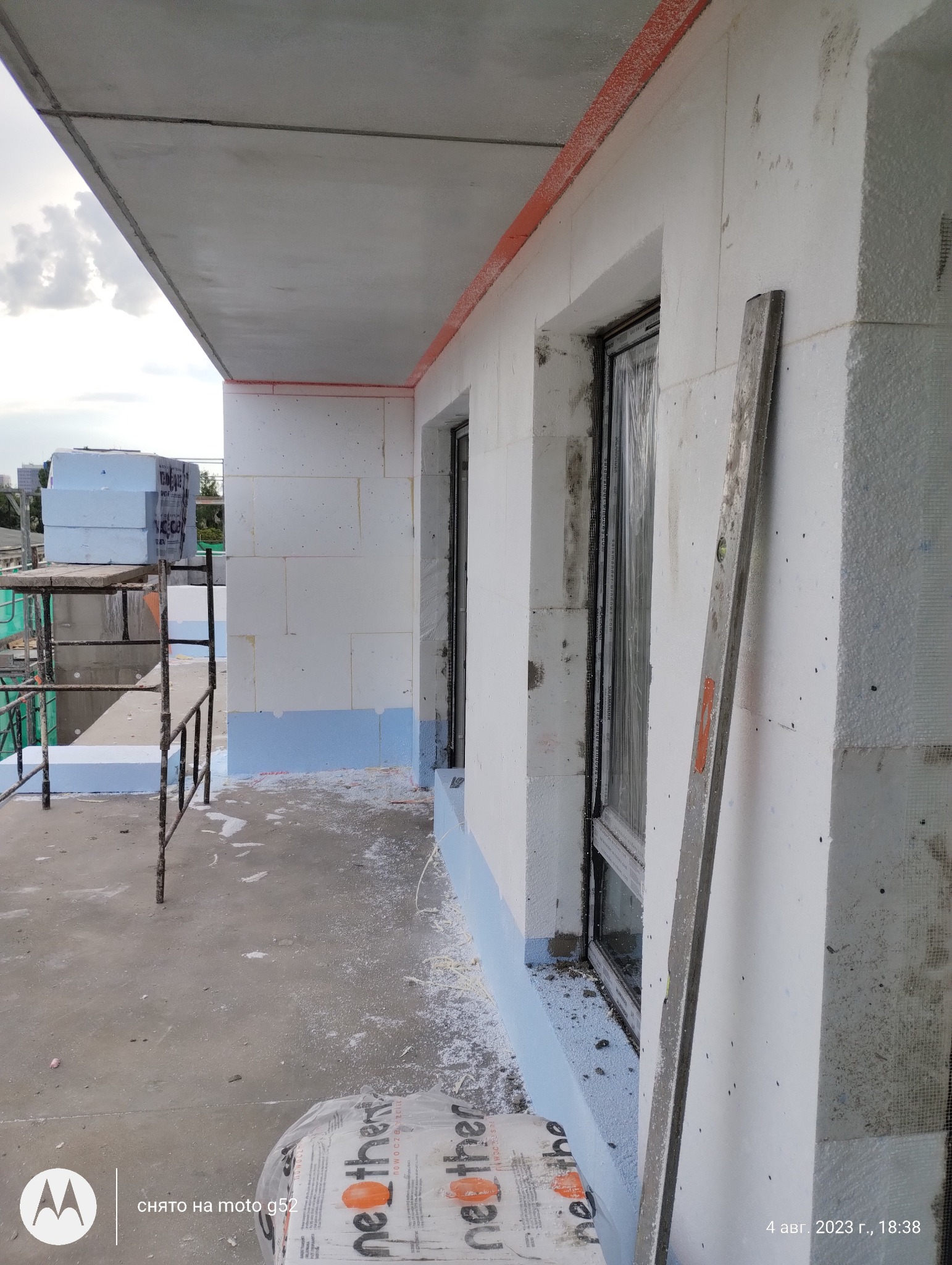 Budynek w trakcie ocieplania elewacji, widoczne białe płyty styropianowe z siatką, okna zabezpieczone folią, poziomica oparta o ścianę, resztki styropianu na podłożu, na podłodze leży paczka...