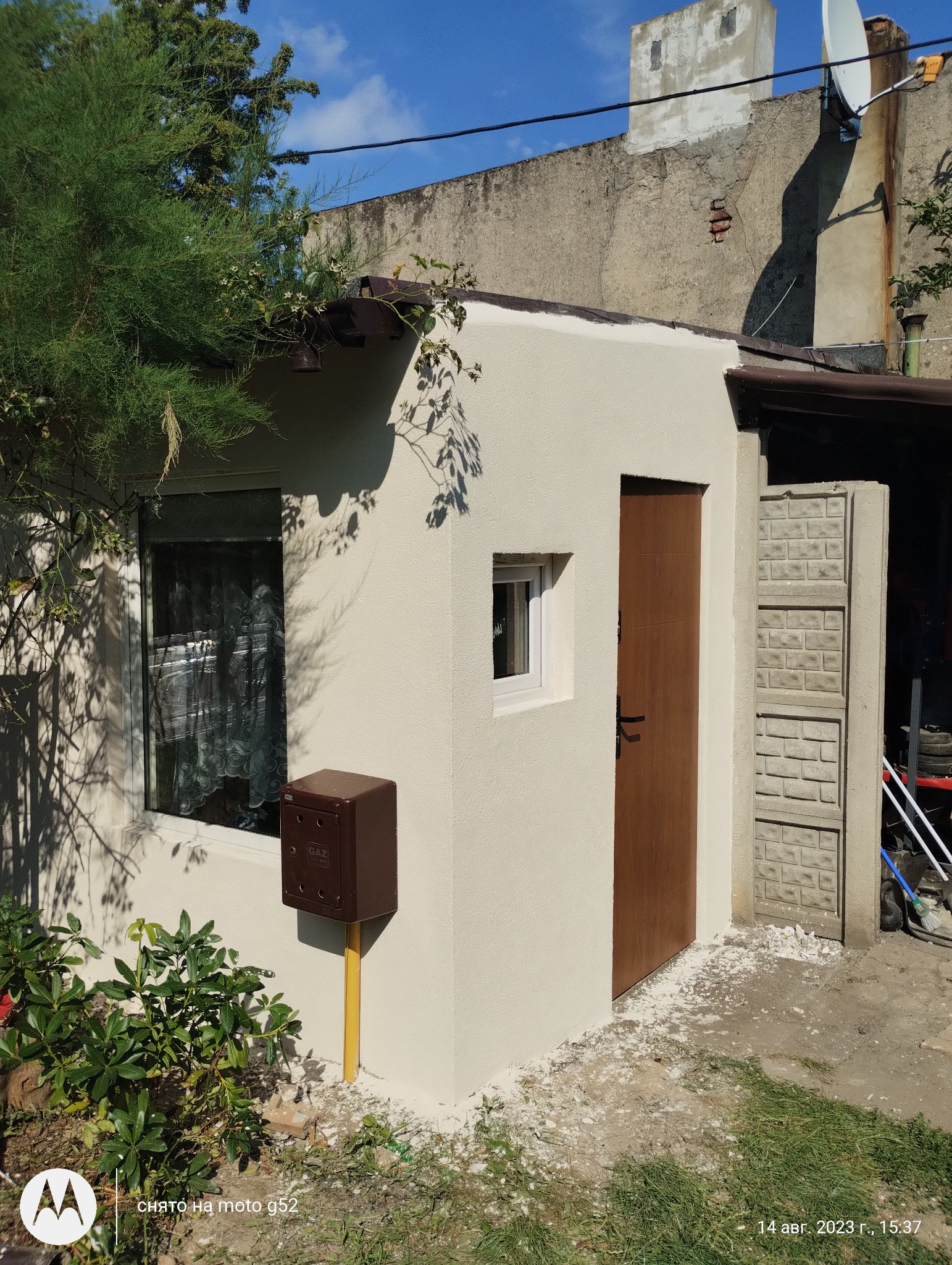 Świeżo otynkowana elewacja budynku z brązową skrzynką gazową, widoczne okno z firanką, drewniane drzwi, betonowe panele i roślinność.