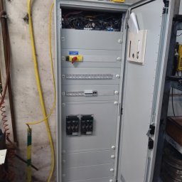 E .L .E .S . Instalacje elektryczne - Otwarta szafa elektryczna z widocznymi bezpiecznikami, wyłącznikiem głównym i przewodami. Żółty kabel biegnie wzdłuż ściany. Widoczne elementy instalacji elektrycznej.