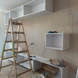 E .L .E .S . Instalacje elektryczne - Montaż białych szafek na ścianie w trakcie remontu. Widoczna drabina i narzędzia. Fragment instalacji elektrycznej.