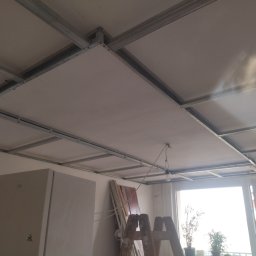 E .L .E .S . Instalacje elektryczne - Montaż sufitu podwieszanego z płyt gipsowych na metalowej konstrukcji. Widoczna drabina, okno i częściowo zakryta szafa. W tle jasne światło dzienne.