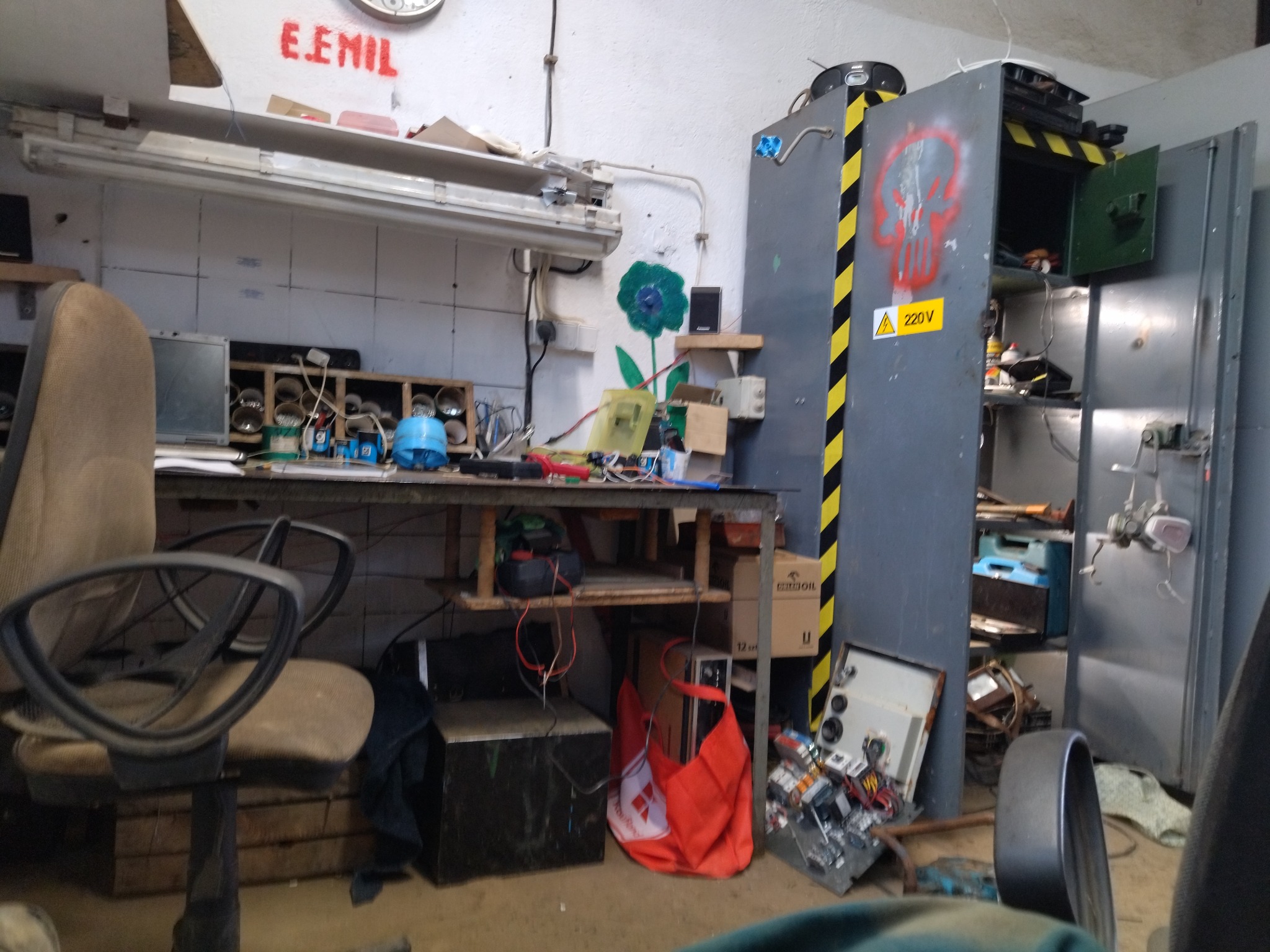 Warsztat elektryczny z biurkiem pełnym narzędzi i szafą z symbolem 220V. Widoczny bałagan i elementy instalacji. Ściana z napisem 'E.EMIL'.