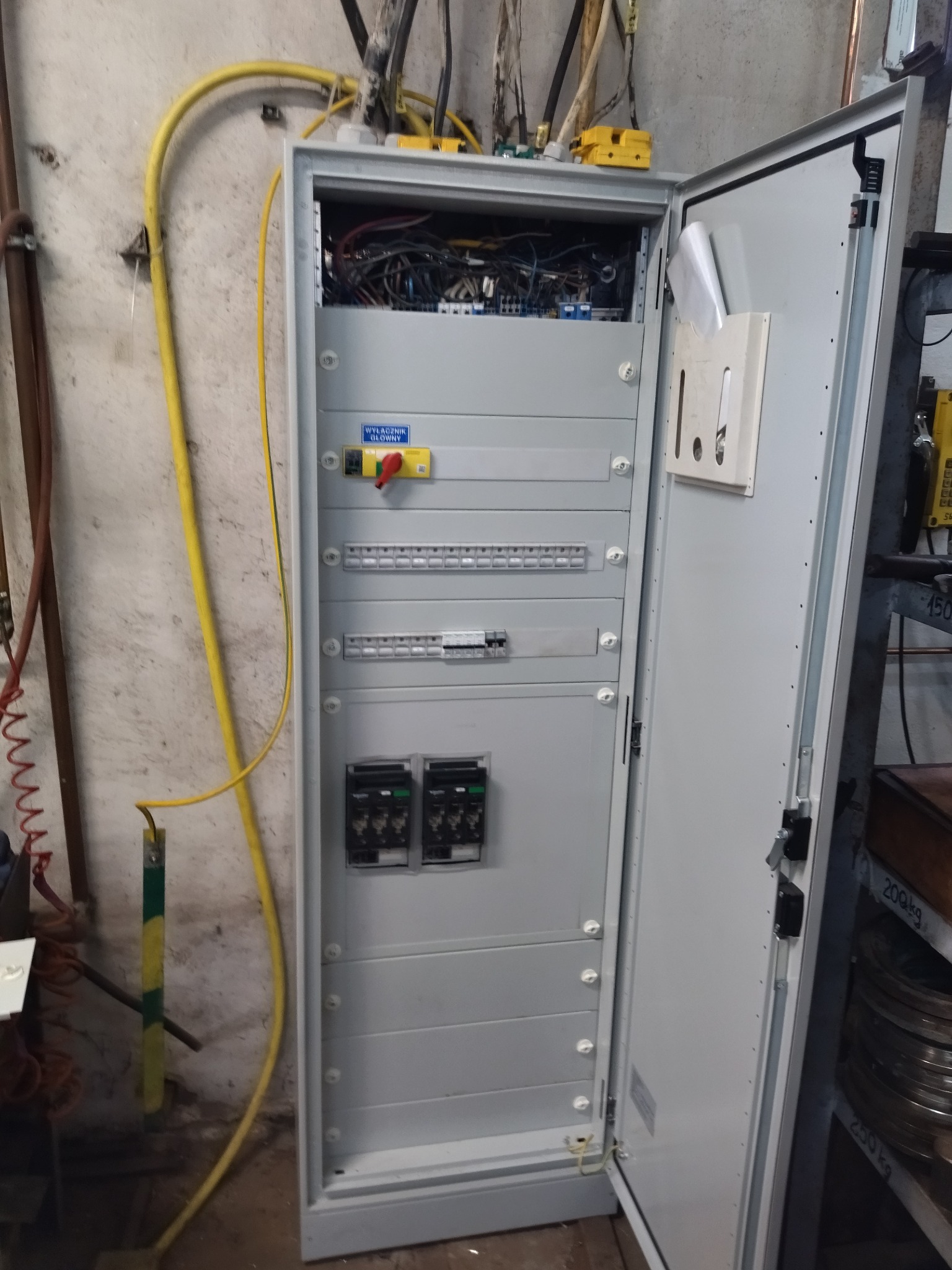 Otwarta szafa elektryczna z widocznymi bezpiecznikami, wyłącznikiem głównym i przewodami. Żółty kabel biegnie wzdłuż ściany. Widoczne elementy instalacji elektrycznej.