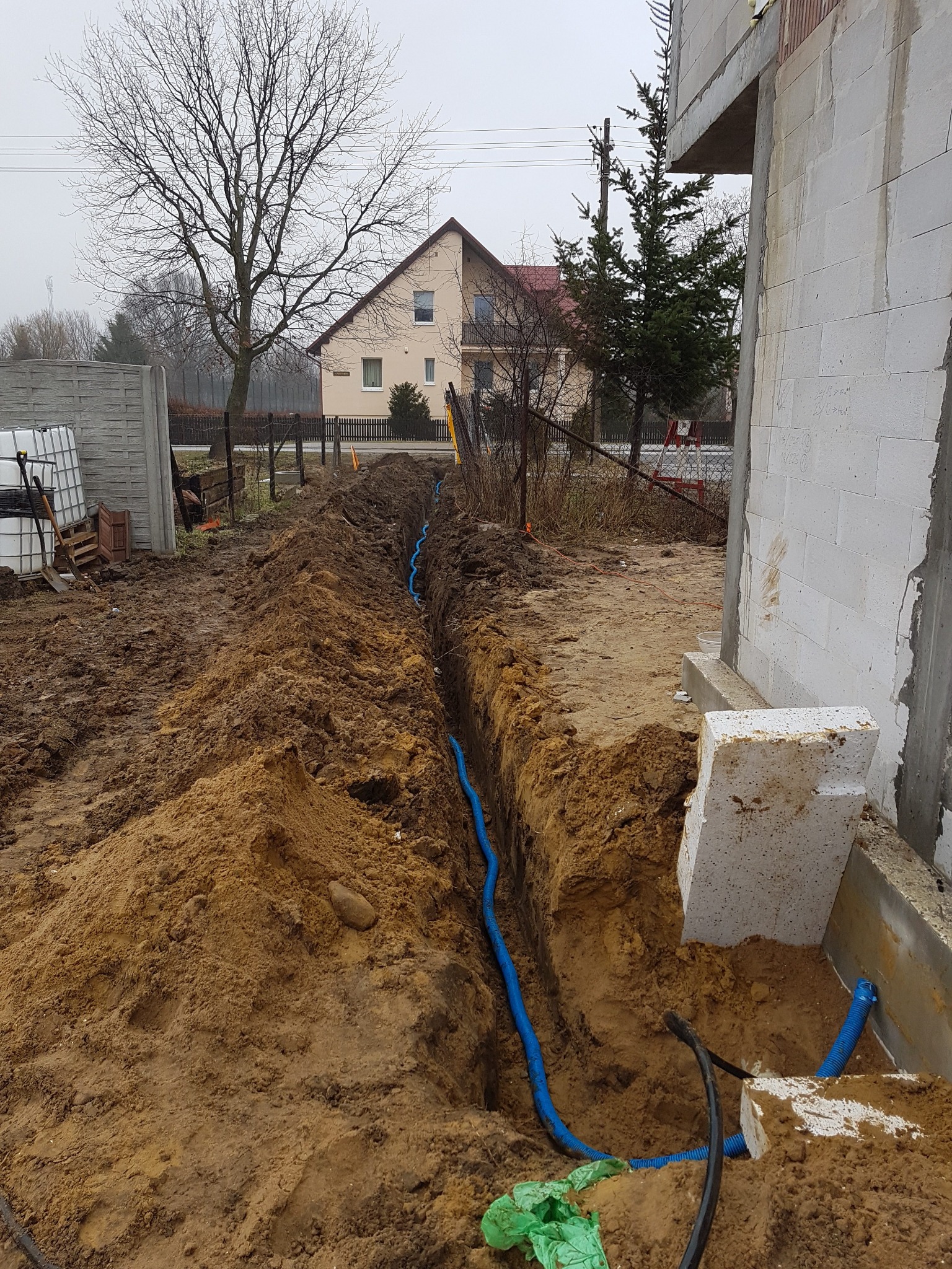 Wykop z ułożonymi niebieskimi rurami kablowymi, prawdopodobnie pod instalacje elektryczne, na terenie budowy domu jednorodzinnego w pochmurny dzień.