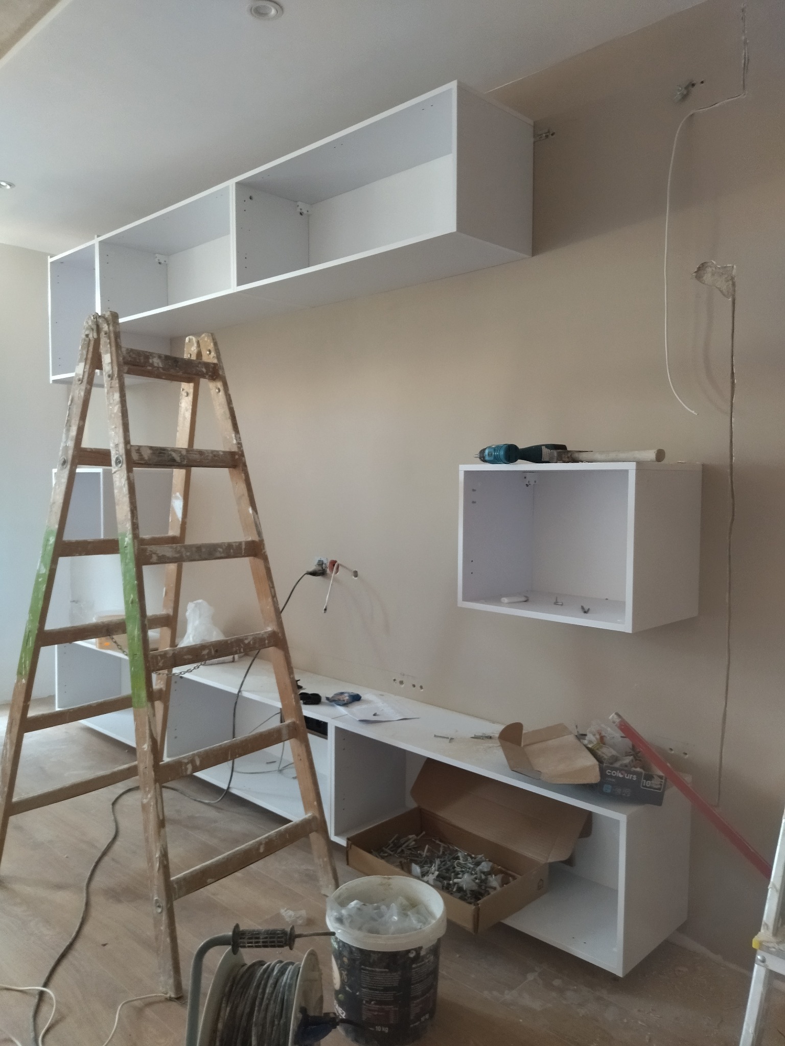 Montaż białych szafek na ścianie w trakcie remontu. Widoczna drabina i narzędzia. Fragment instalacji elektrycznej.