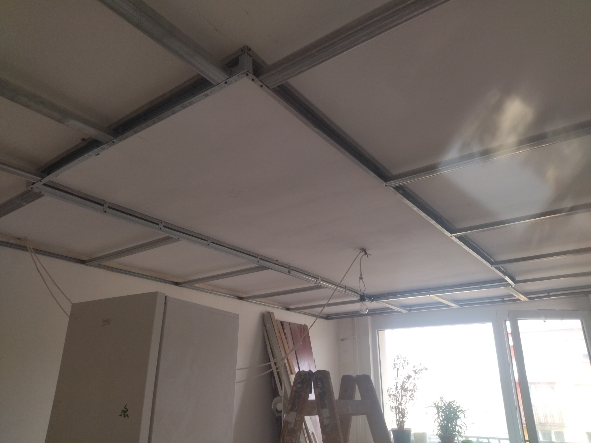 Montaż sufitu podwieszanego z płyt gipsowych na metalowej konstrukcji. Widoczna drabina, okno i częściowo zakryta szafa. W tle jasne światło dzienne.