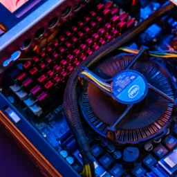 Wnętrze komputera stacjonarnego z widoczną płytą gł&oacute;wną, radiatorem z czerwonymi elementami i wentylatorem Intel, oświetlone niebieskim światłem.