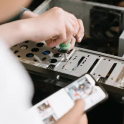 Osoba przykręca śrubkę wewnątrz obudowy komputera, trzymając w drugiej ręce smartfon z otwartą instrukcją lub dokumentacją techniczną.