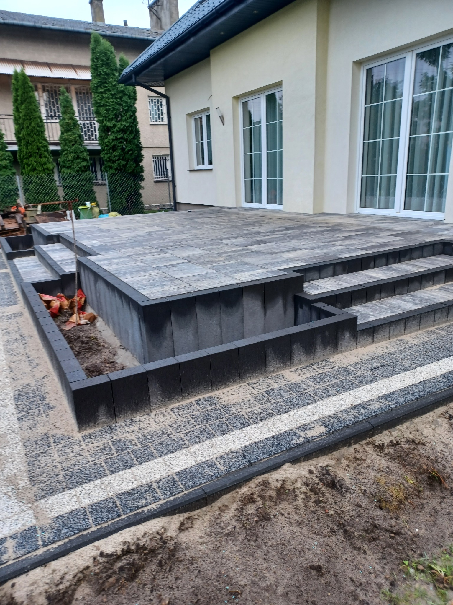 Nowoczesny taras z kostki brukowej w odcieniach szarości, z niskimi schodami i obrzeżem, świeżo po ułożeniu, z widocznymi resztkami piasku na powierzchni i przygotowanym miejscem na rośliny.