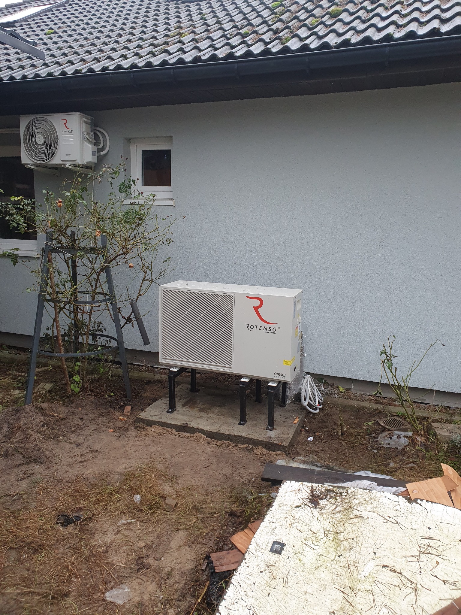 Zewnętrzna jednostka pompy ciepła Rotenso zamontowana na betonowej podstawie i metalowych wspornikach, z widoczną jednostką wewnętrzną na ścianie budynku z szarą elewacją i ciemną dachówką, obok...