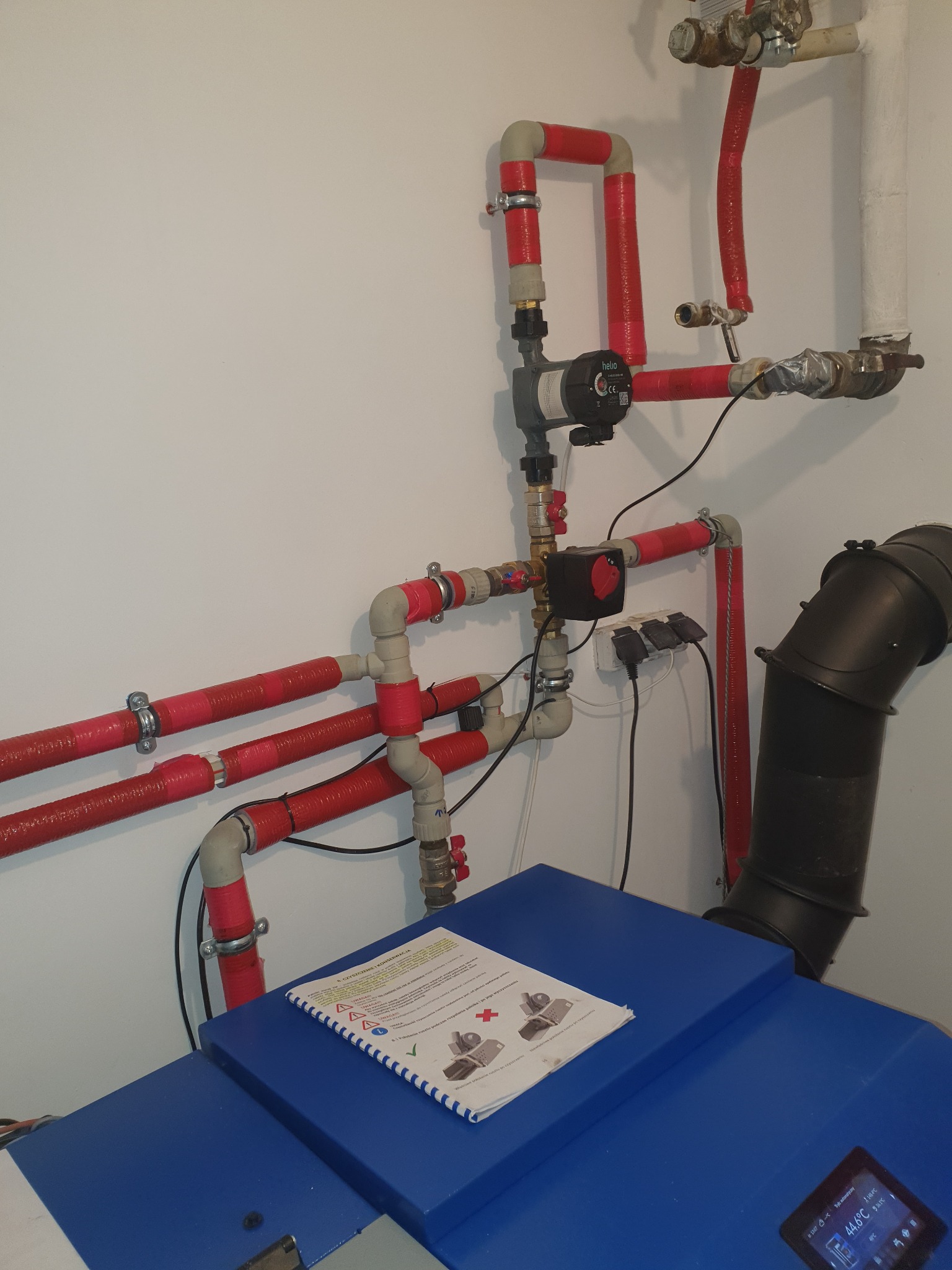 Fragment instalacji grzewczej z rurami owiniętymi czerwoną taśmą izolacyjną, widoczny piec z wyświetlaczem i instrukcja obsługi na wierzchu.