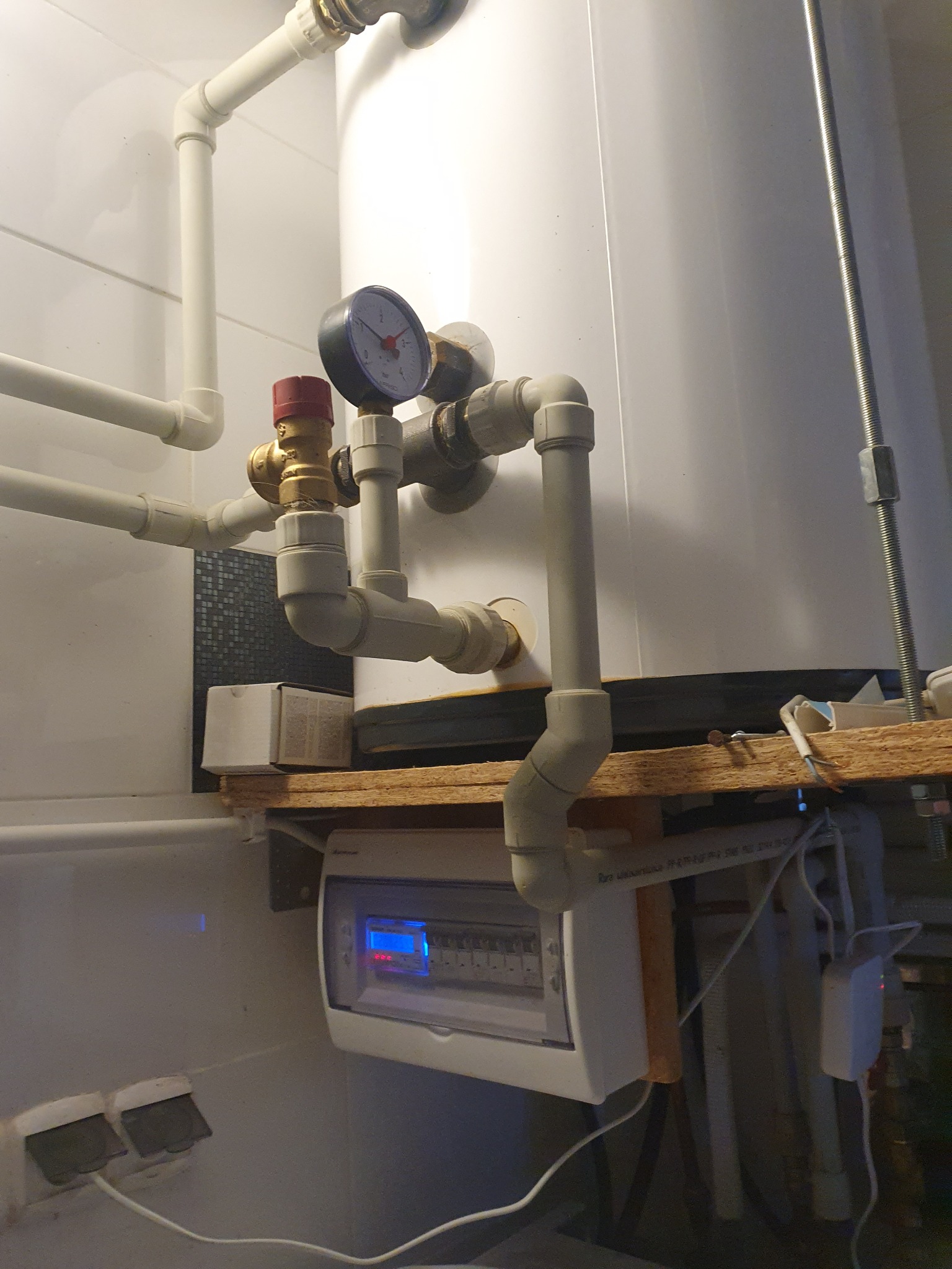 Bojler z podłączonymi rurami z tworzywa, manometrem i zaworem bezpieczeństwa, zamontowany na drewnianej półce nad skrzynką bezpiecznikową w pomieszczeniu z białymi płytkami.