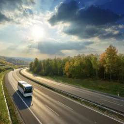 Biały autokar na zakręcie autostrady w jesiennej scenerii, promienie słoneczne przebijają się przez chmury.