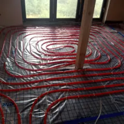 Czerwone rury ogrzewania podłogowego ułożone spiralnie na folii z rastrem, widoczne okno i surowa ściana z cegły.