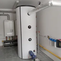 Biały, cylindryczny zbiornik akumulacyjny z czarnymi okrągłymi zaślepkami, podłączony do instalacji, obok wiszący piec gazowy i filtry wody, w pomieszczeniu technicznym z szarą podłogą.
