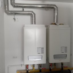 TECH INSTALL TECHNIKA GRZEWCZA I SANITARNA Błażej Śmigiel - Dwa białe, wiszące kotły gazowe marki Immergas z podłączonymi rurami odprowadzającymi spaliny, zamontowane na białej ścianie w nowym pomieszczeniu z widocznymi gniazdkami elektrycznymi i rurami...