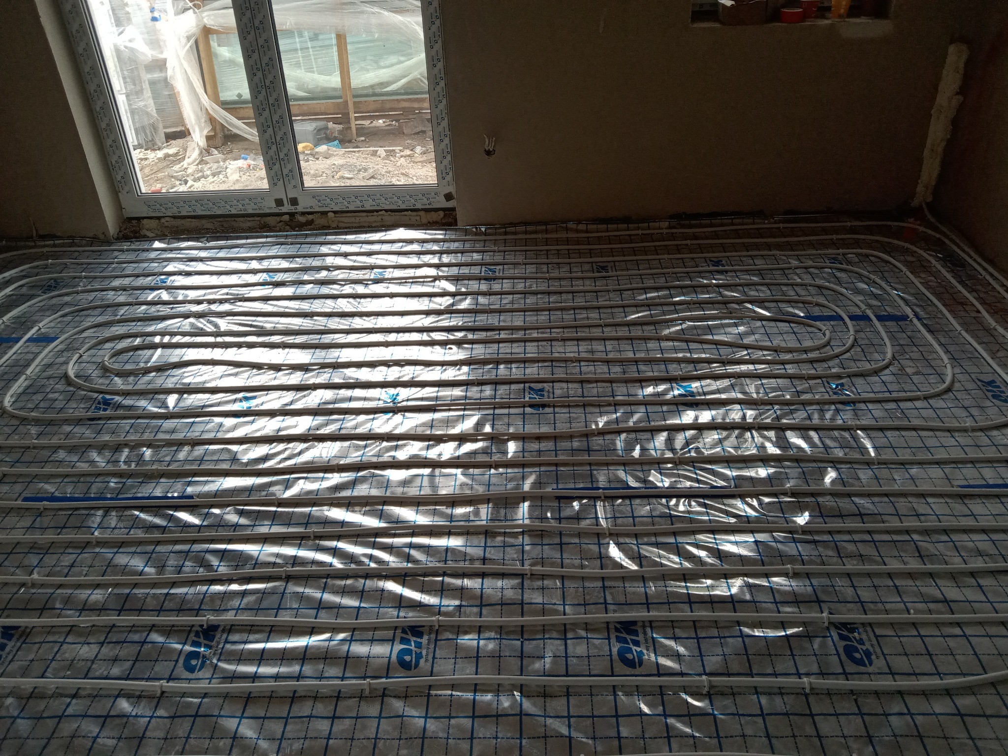 Białe rurki ogrzewania podłogowego ułożone na folii z niebieską siatką, widoczne okno z zewnątrz oraz fragment ściany w surowym stanie.